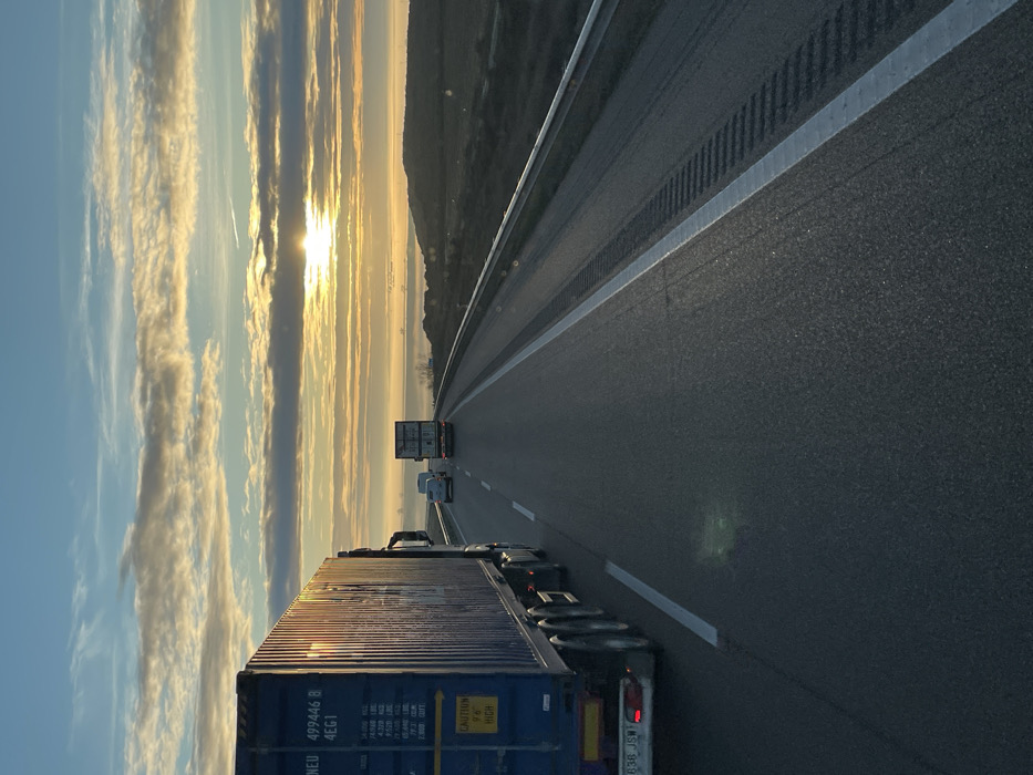 A highway stretches forward under a vibrant sky, the sun breaking through linear clouds at the horizon. A large semi-truck dominates the foreground, with other vehicles, including a box truck, visible further ahead. The road markings are clearly defined, with reflective paint catching the light. A metallic guardrail runs along the side, and a muted, hilly landscape sits just beyond the highway. The perspective emphasizes the length and openness of the road.