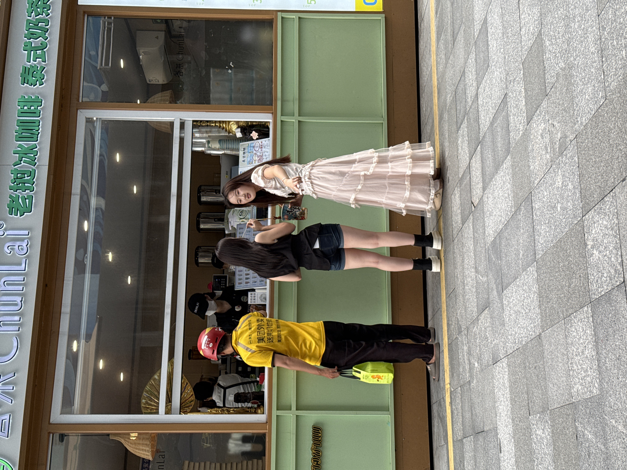 In Huizhou, China, at a street-facing beverage kiosk, three individuals are visible. To the left, a male delivery person wearing a yellow shirt and black pants, with a red helmet, holds a bright yellow bag. In the center, a female with long dark hair and denim shorts stands. To her right, another female in a long, layered pink dress is interacting with the first female. Behind them, inside the kiosk, two employees are present, one wearing a black cap and mask. The kiosk features green paneling and a large window displaying beverages. The scene takes place on a paved sidewalk with a yellow line marking the edge.