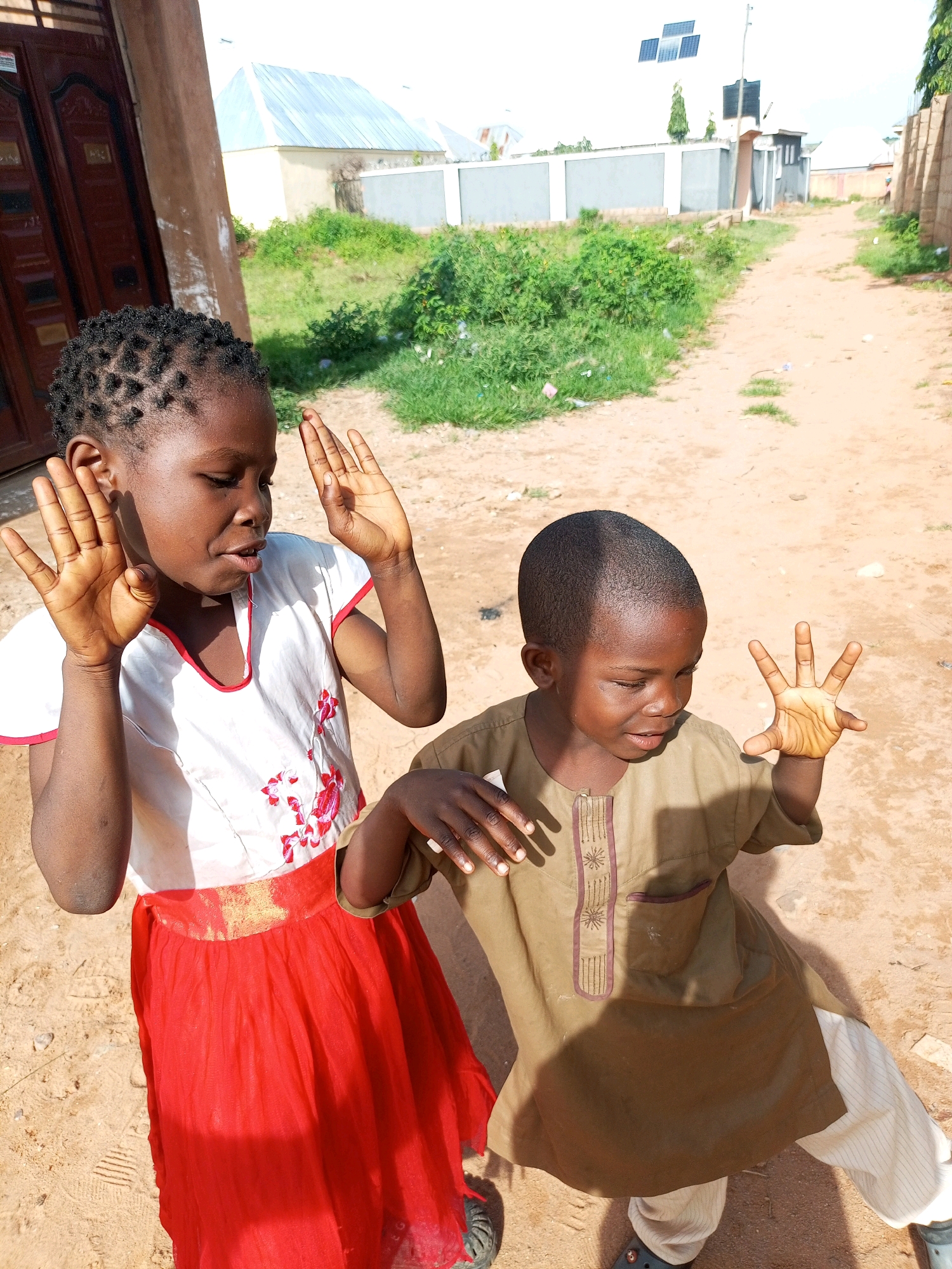 In Tempure, Nigeria, two young children, a girl and a boy, stand on a dusty, unpaved road. The girl, on the left, is wearing a white top with red trim and a vibrant red skirt. Her hands are raised, palms out, as if gesturing. The boy, on the right, is dressed in a light brown tunic with decorative stitching and light-colored striped trousers. His right hand is also raised, fingers spread. The background reveals a building with a corrugated metal roof, some greenery, and a fence, suggesting a residential area. The scene appears to be a candid shot captured outdoors during daylight hours.