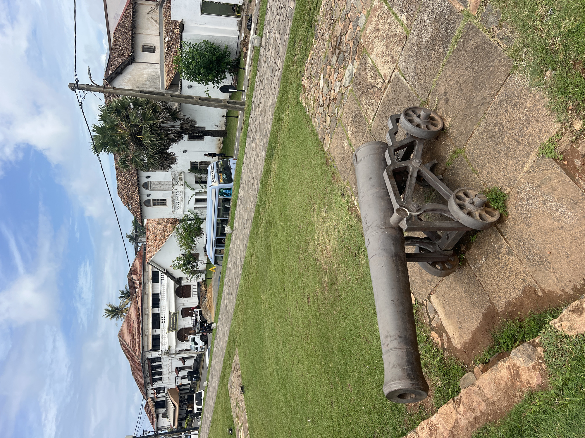 In Galle, Sri Lanka, a historic cannon rests on a stone platform in front of a manicured lawn. The cannon, made of dark metal, is mounted on a wheeled carriage. In the background, a street lines the edge of the lawn, with a row of traditional white buildings featuring terracotta tiled roofs. A blue bus with the word "Journey" visible on its side is driving on the street, and a white truck is parked near one of the buildings. Palm trees and other greenery are scattered throughout the scene, and the sky is partially cloudy.