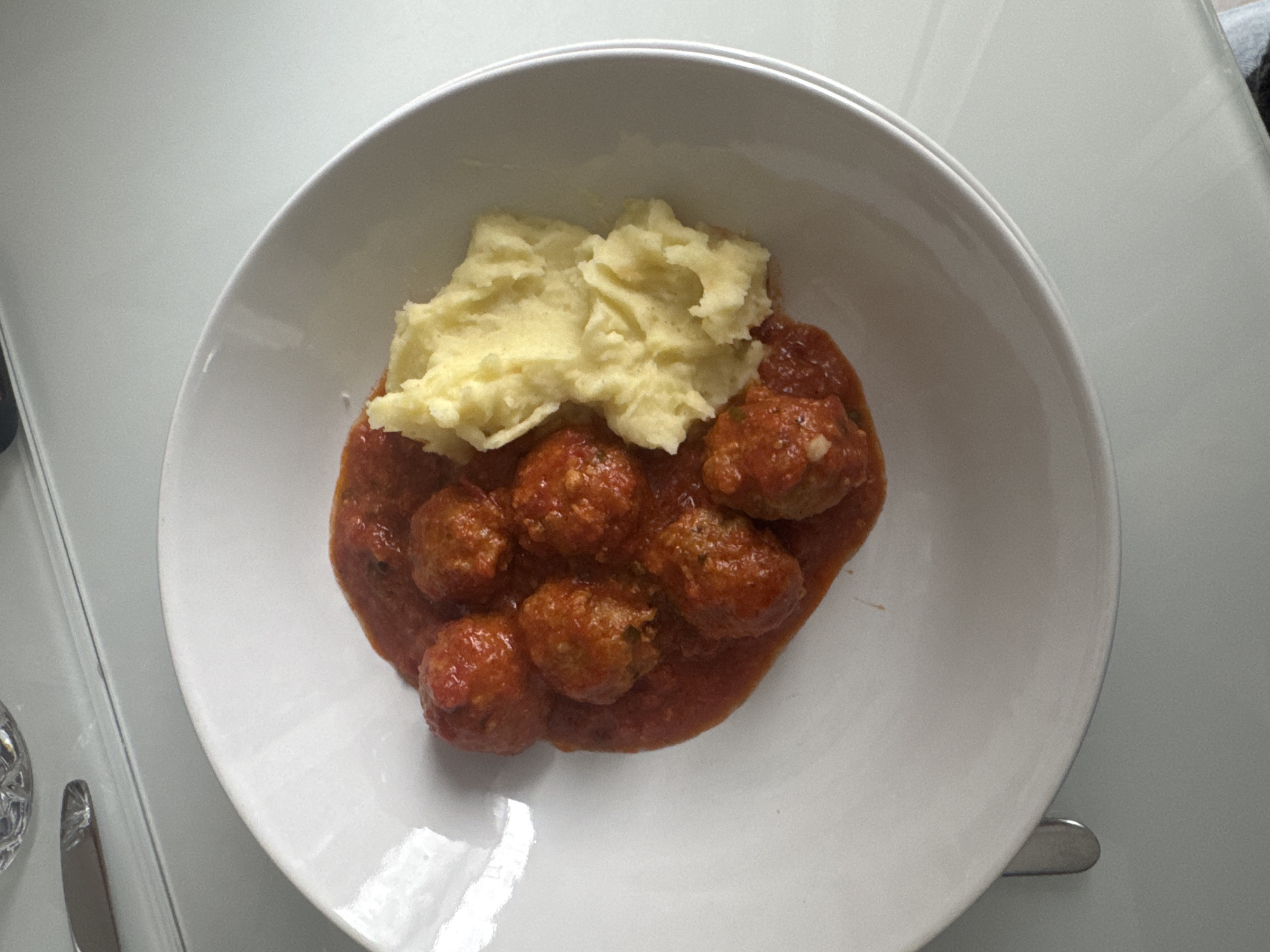 A top-down view of a white ceramic bowl containing meatballs in tomato sauce and a serving of mashed potatoes. The bowl is placed on a white, reflective surface, likely a table. The scene appears to be indoors, with a blurred glimpse of cutlery and possibly other objects on the left side of the frame. There is no indication of the image being taken in Milan, Italy.