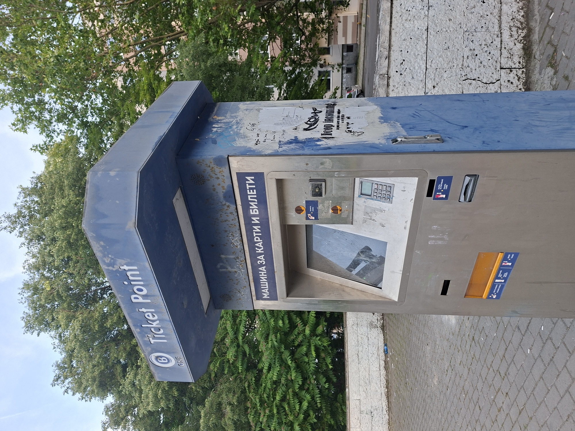 An outdoor ticket vending machine, exhibiting significant wear and visible damage, is centrally positioned on a grey paved surface in Varna, Bulgaria. The machine features a two-tone design, with a weathered blue canopy atop a predominantly silver metallic body. The blue canopy bears the text "Ticket Point" in white lettering, accompanied by a circular logo enclosing a 'B' and the word "ONE". Rust and discoloration are apparent on the blue surfaces.
The silver operational face of the machine displays "МАШИНА ЗА КАРТИ И БИЛЕТИ" (MACHINE FOR CARDS AND TICKETS) in a blue strip at the top. Below this, a large rectangular screen is severely cracked and non-functional. To the right of the damaged screen, multiple slots and icons are present, including one suggesting card insertion. A numeric keypad is located below the screen, showing digits 0-9. Further down, a slot is labeled "КАРТА ЗА ПЪТУВАНЕ" (TRAVEL CARD) and "TRAVEL CARD". At the bottom, a horizontal black slot is visible above a larger orange dispensing tray labeled with icons for "ВЪВЕДЕТЕ СУМА / CHANGE GIVEN", "ФИСКАЛЕН БОН / FISCAL RECEIPT", and "БИЛЕТ / TICKET" (TICKET).
The blue side panels of the machine are heavily soiled, marked with adhesive residue from removed posters, and faint graffiti, including the visible phrase "NOW drop incoming" on a torn fragment. A metal handle is affixed to the right side panel.
The ground around the machine is paved with grey interlocking concrete blocks. To the left and in the immediate background, dense green foliage from mature trees is prominent. To the right, beyond a low, textured white stone wall exhibiting a vertical crack, additional trees partially obscure a light-colored building and a parked white car on an asphalt road. Litter, including a blue plastic bottle and cigarette butts, is scattered on the paved surface near the base of the wall. The scene is illuminated by ambient daylight.