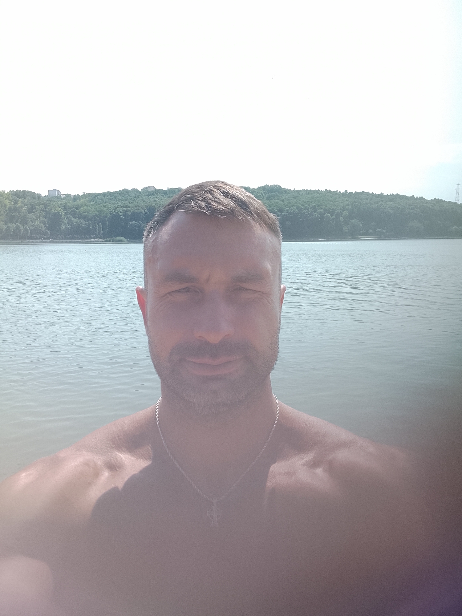 A man is taking a selfie in front of a body of water, likely a lake or river, on a sunny day. He is shirtless, wearing a silver chain with a cross pendant. The background shows a treeline with some buildings and a power line tower in the distance, suggesting an urban or suburban setting near nature. The image does not provide enough information to identify the specific location in Chișinău, Moldova.