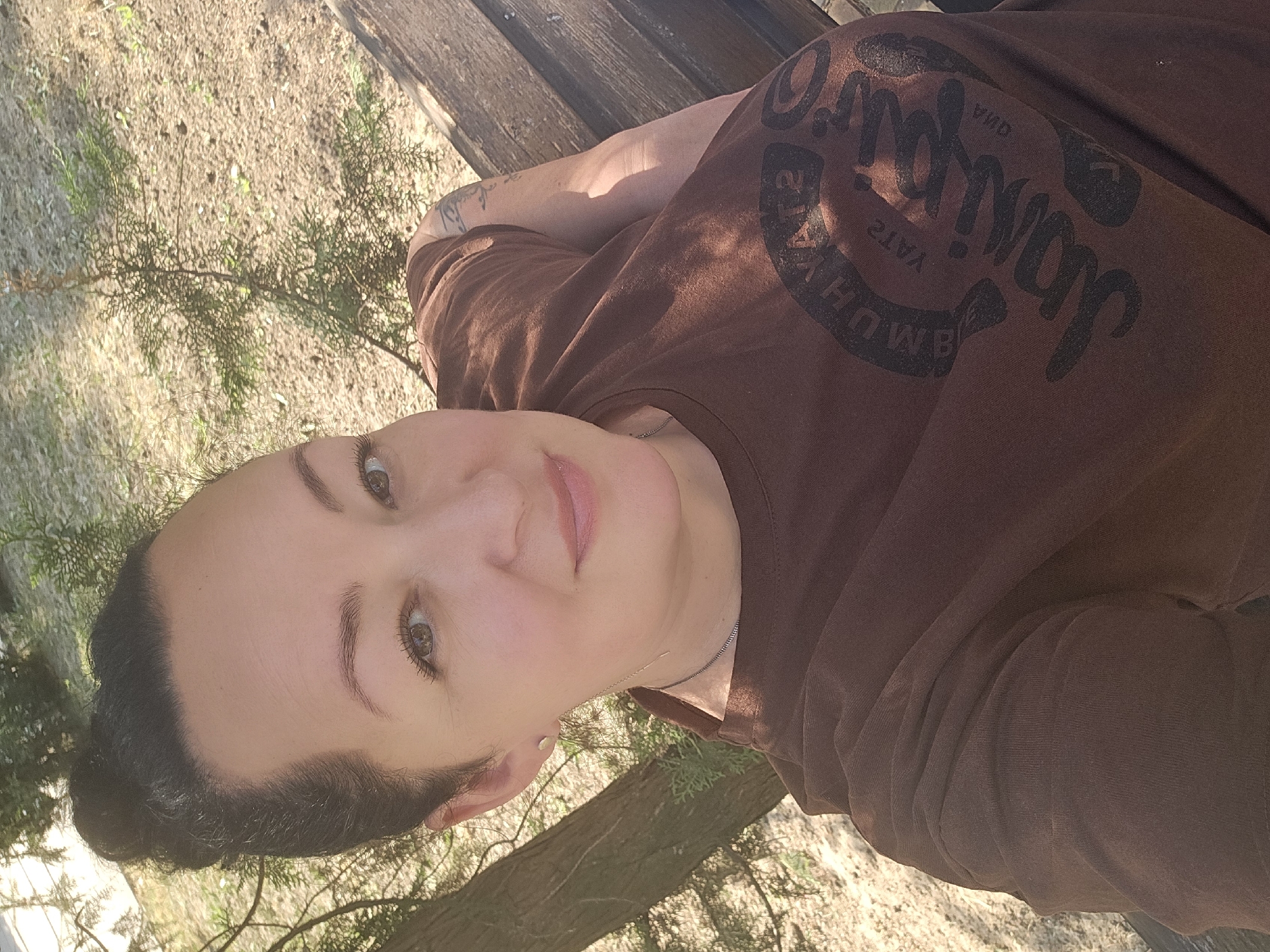 A woman is taking a selfie outdoors, seated on a wooden bench. She has her dark hair pulled up in a bun and is wearing a brown t-shirt with a graphic design on it, visible on the chest and shoulder. The background consists of trees and sandy ground. The setting appears to be a park or natural area.