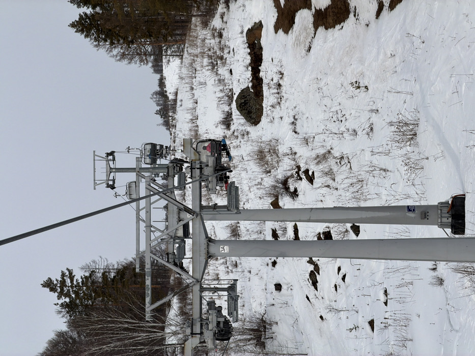 The scene depicts a ski slope in Novoabzakovo, Russia, under an overcast sky. A multi-column grey metal chairlift pylon, marked "3A," stands prominently in the foreground. Overhead, a steel cable supports several grey chairlifts. One chairlift in the mid-ground carries an individual clad in a pink jacket and blue pants, with a snowboard attached to their feet, ascending the slope. Other empty chairlifts are visible both behind and further up the line. The ground is partially covered in snow, revealing patches of dry, brown vegetation and scattered dark rocks. Visible ski tracks traverse the snowy surface. To the left, a cluster of bare deciduous trees stands against a backdrop of darker coniferous trees. To the right, the slope continues upwards, bordered by more coniferous trees along the horizon. Patches of exposed brown earth or mud are visible on the far right side of the image.