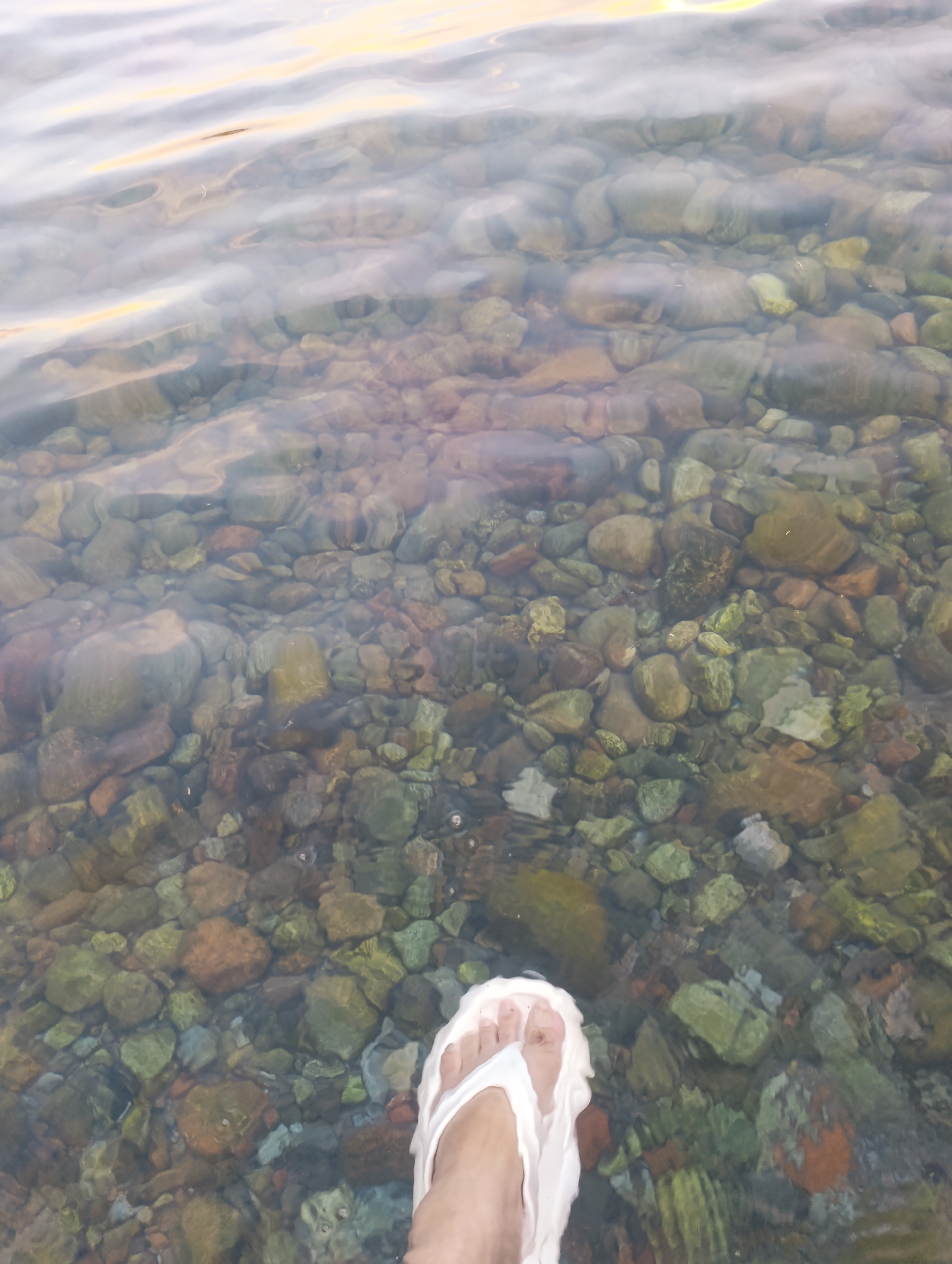 A person's foot, wearing a white flip-flop, is submerged in shallow, clear water. The water reveals a bed of smooth, rounded pebbles of various sizes and colors, predominantly brown, green, and gray. Gentle ripples on the water's surface catch the light, creating soft reflections. The background is out of focus, showing a blurry expanse of water with a warm, diffused light, possibly from the sky. This scene appears to be a natural waterfront, likely a shallow river or lake shore, within the vicinity of Bayet, Kyrgyzstan.