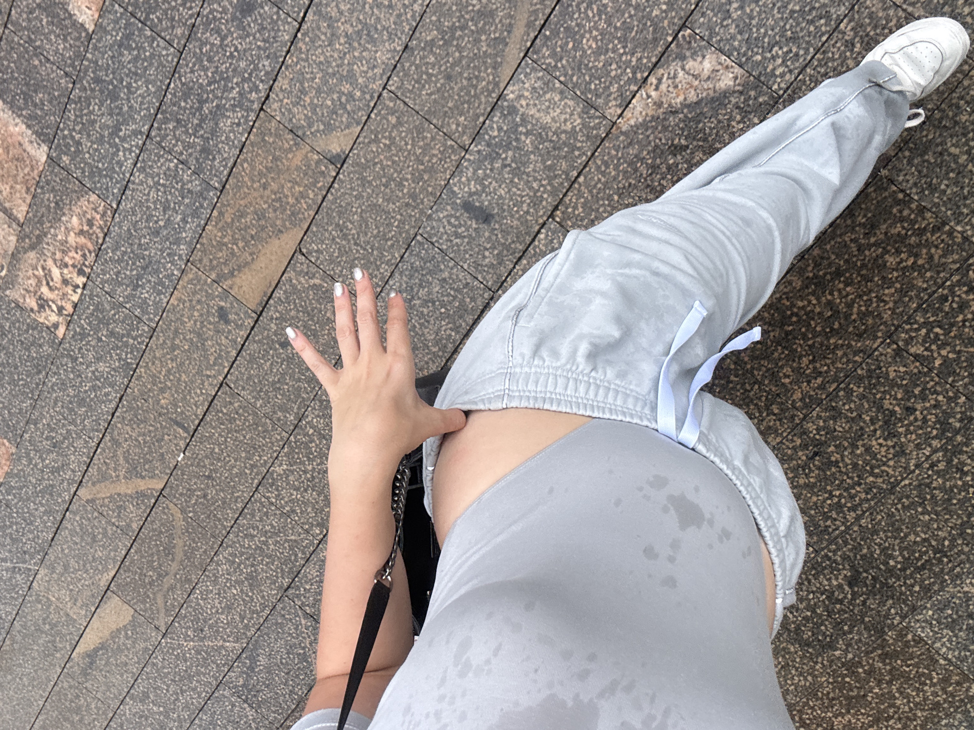 A person's torso and legs are shown from a high angle, standing on a paved walkway. The walkway is made of rectangular stone tiles arranged diagonally. The person is wearing a light gray sports bra or crop top that appears to be damp with sweat, and light gray sweatpants with a drawstring closure. Their left arm is extended, with the hand with silver nail polish reaching towards their stomach. A black purse with a chain strap is visible over the person's left shoulder. A white sneaker is visible on the person's right foot. The overall scene appears to be outdoors in a public area.