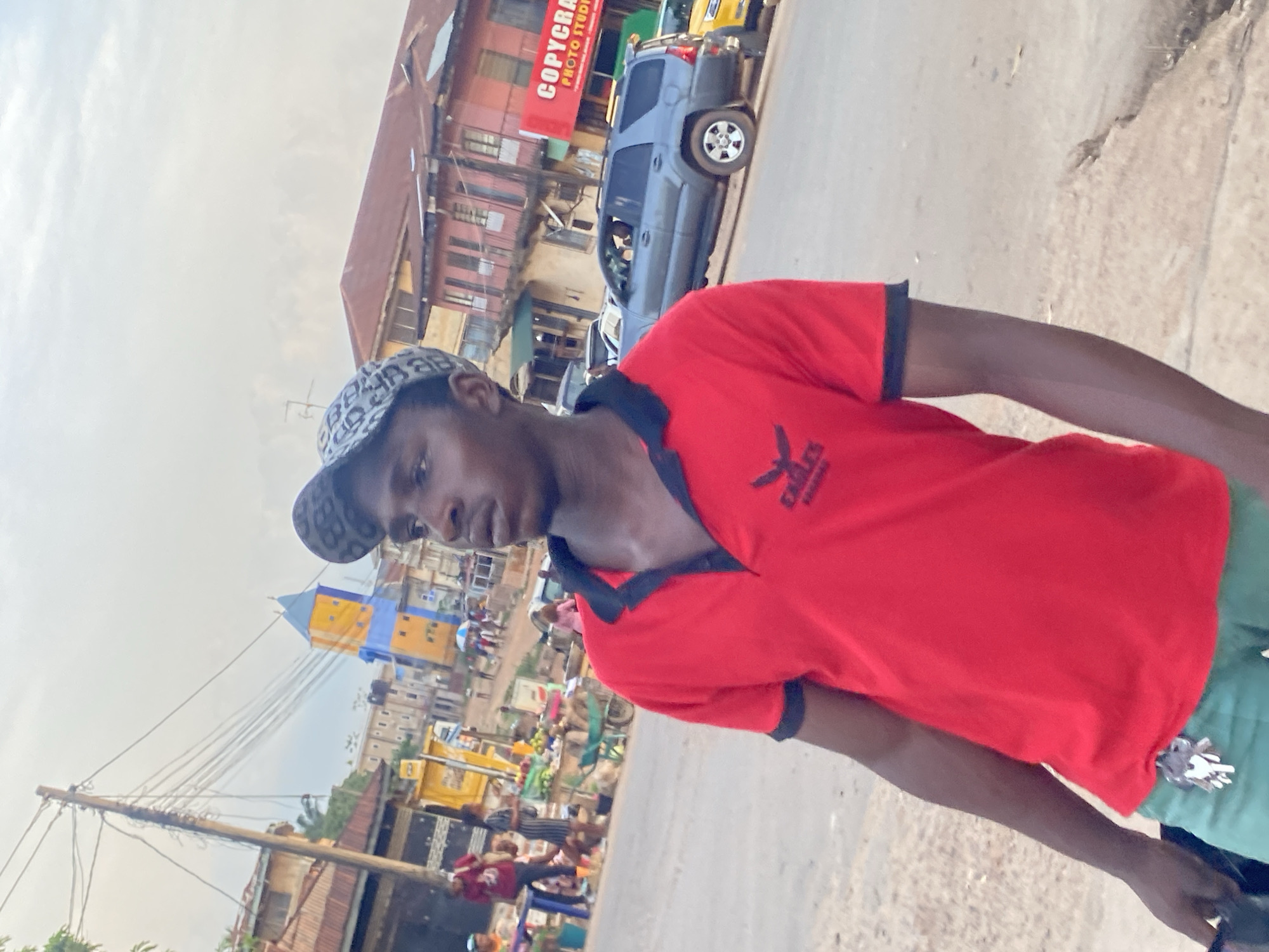 A man in a red shirt and patterned baseball cap stands on a street in Abofifia, Nigeria. Buildings and parked cars line the street. A sign for "COPYCRAFT PHOTO STUDIO" is visible on one of the buildings. A utility pole with wires stretches into the scene. People and market stalls populate the background. The sky is overcast.