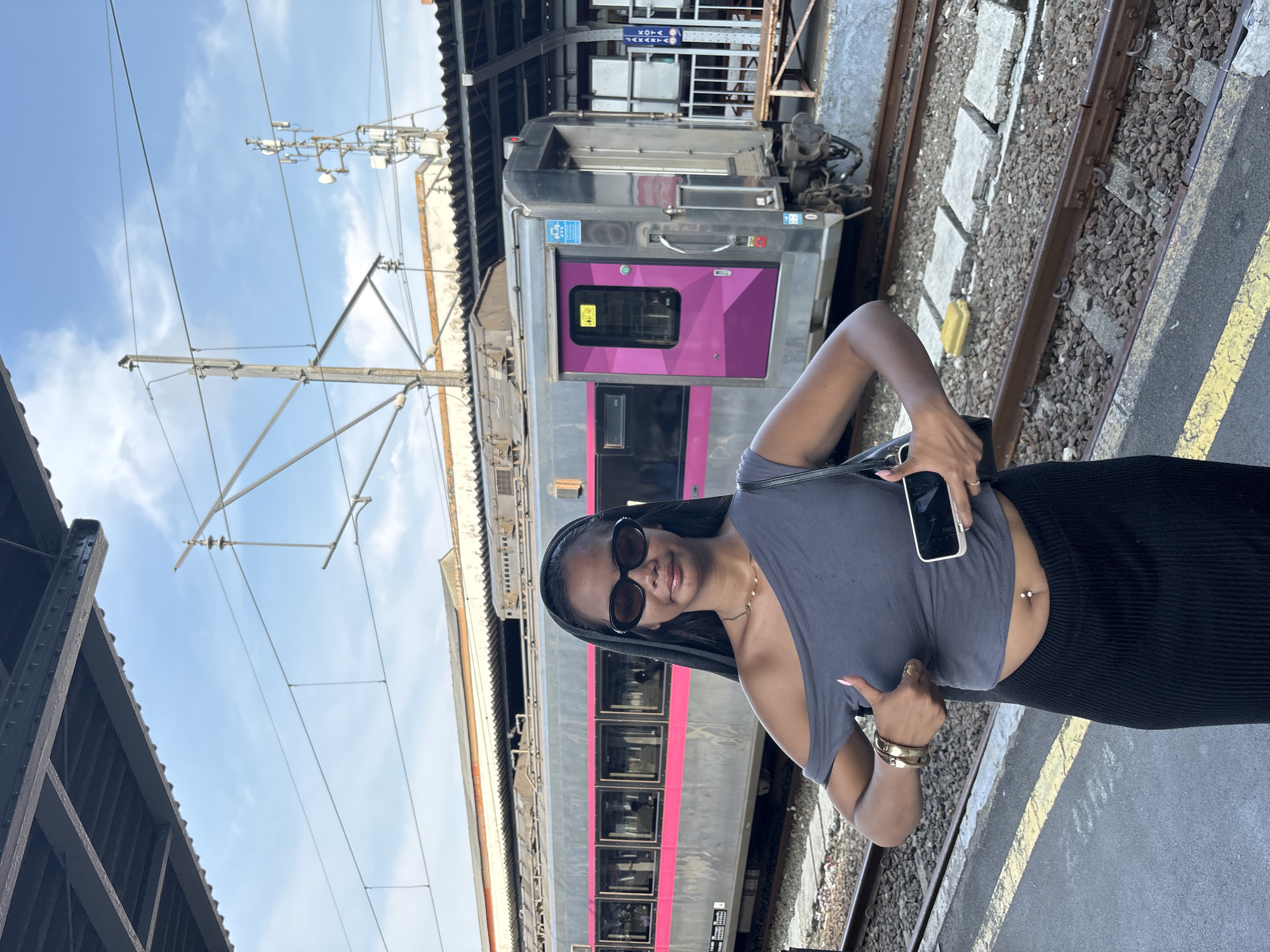 A woman stands on a train platform next to a train with silver cars and pink stripes and door. She is wearing a grey crop top, black pants, sunglasses, and a gold necklace, and she is holding a phone in her left hand and giving a thumbs up with her right hand. Overhead electrical wires are visible above the train. The city location is not discernible from the image.