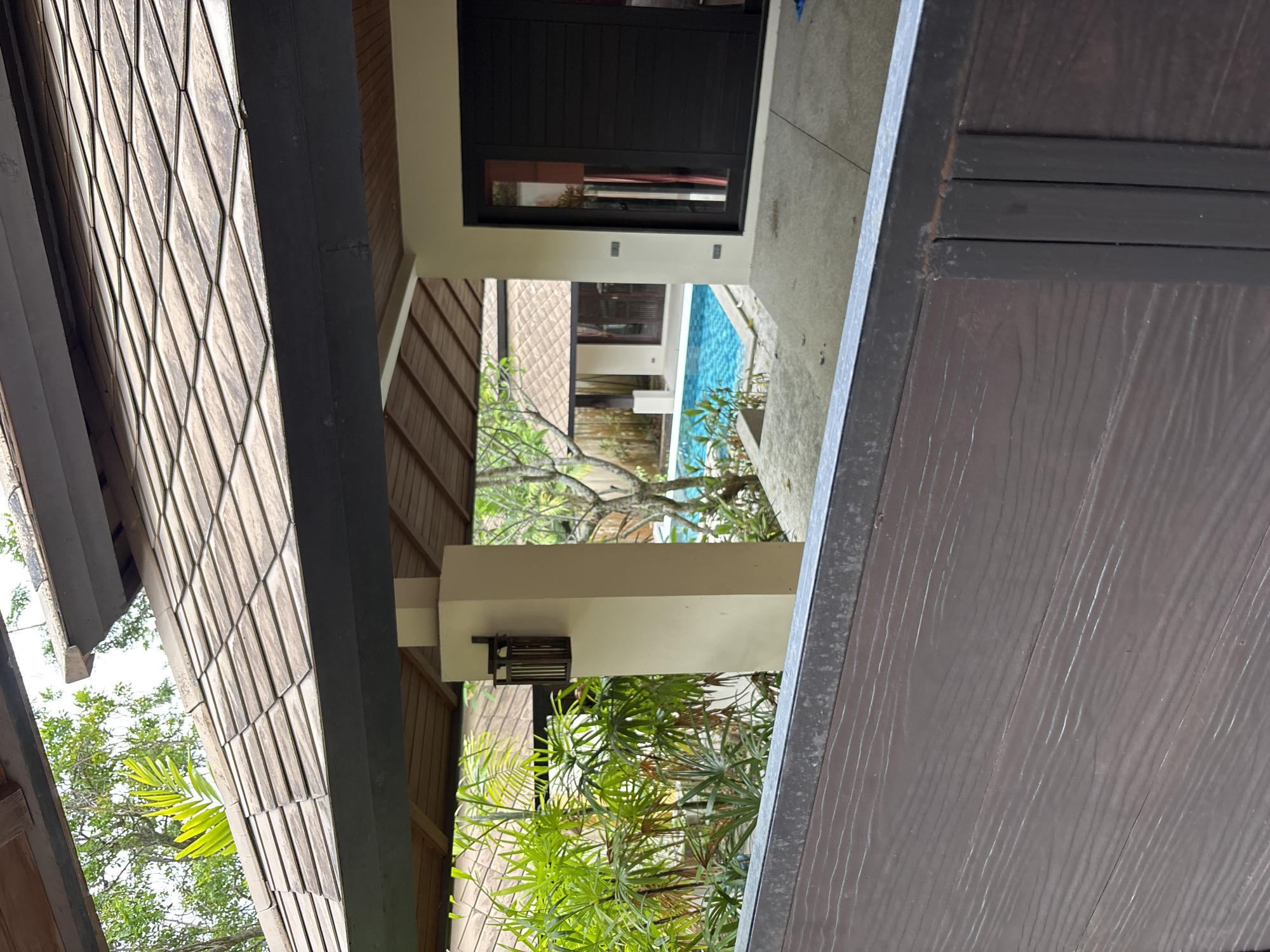 The image shows an exterior view of a building with a tiled roof, supported by a beige pillar with a wall-mounted lantern. In the background, a swimming pool with blue tiles is visible, surrounded by lush green foliage and trees. A modern door with dark panels is present on the right side of the frame. The foreground features a dark brown wooden fence. The scene is set in an outdoor environment, likely a resort or private residence.