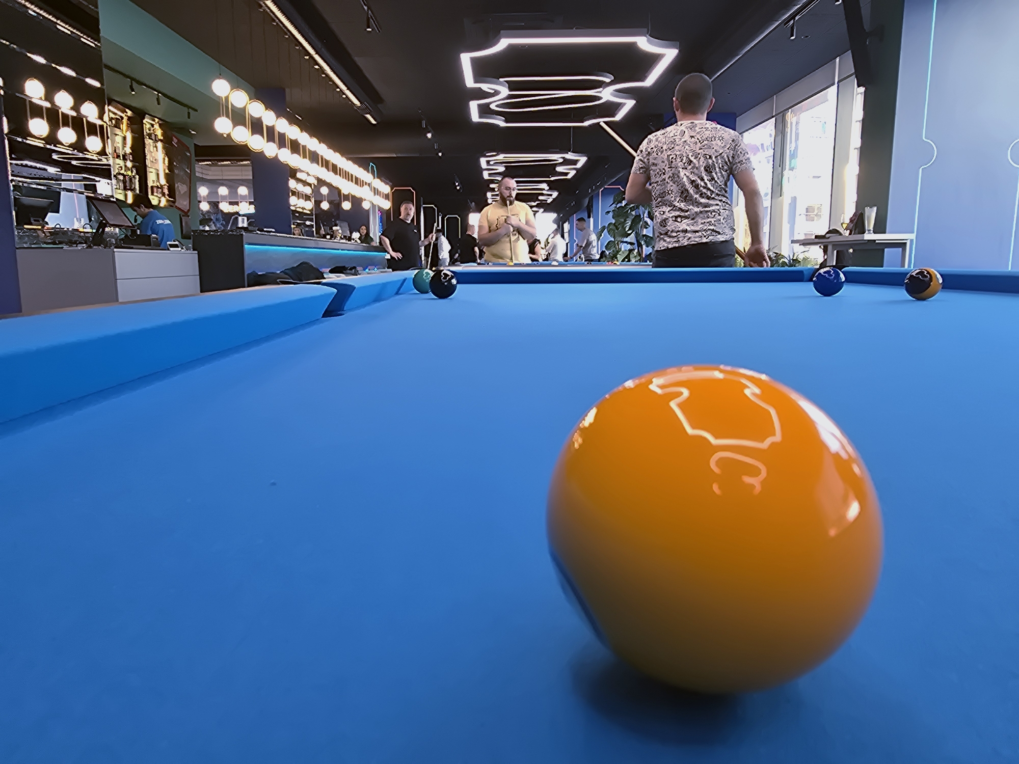 The image shows a view across a blue pool table, with an orange ball prominently in the foreground. In the mid-ground, there are three other pool balls in blue, green, and brown tones, spread across the table. Several individuals are visible, with one person positioned behind the table holding a cue stick as if preparing to strike a ball. Other people stand and sit in the background. Overhead, modern lighting fixtures are present, contributing to the interior ambiance. The scene appears to be inside a recreational or entertainment venue. According to online sources, the location is identified as Play Park Varna, an entertainment venue located in Varna, Bulgaria.