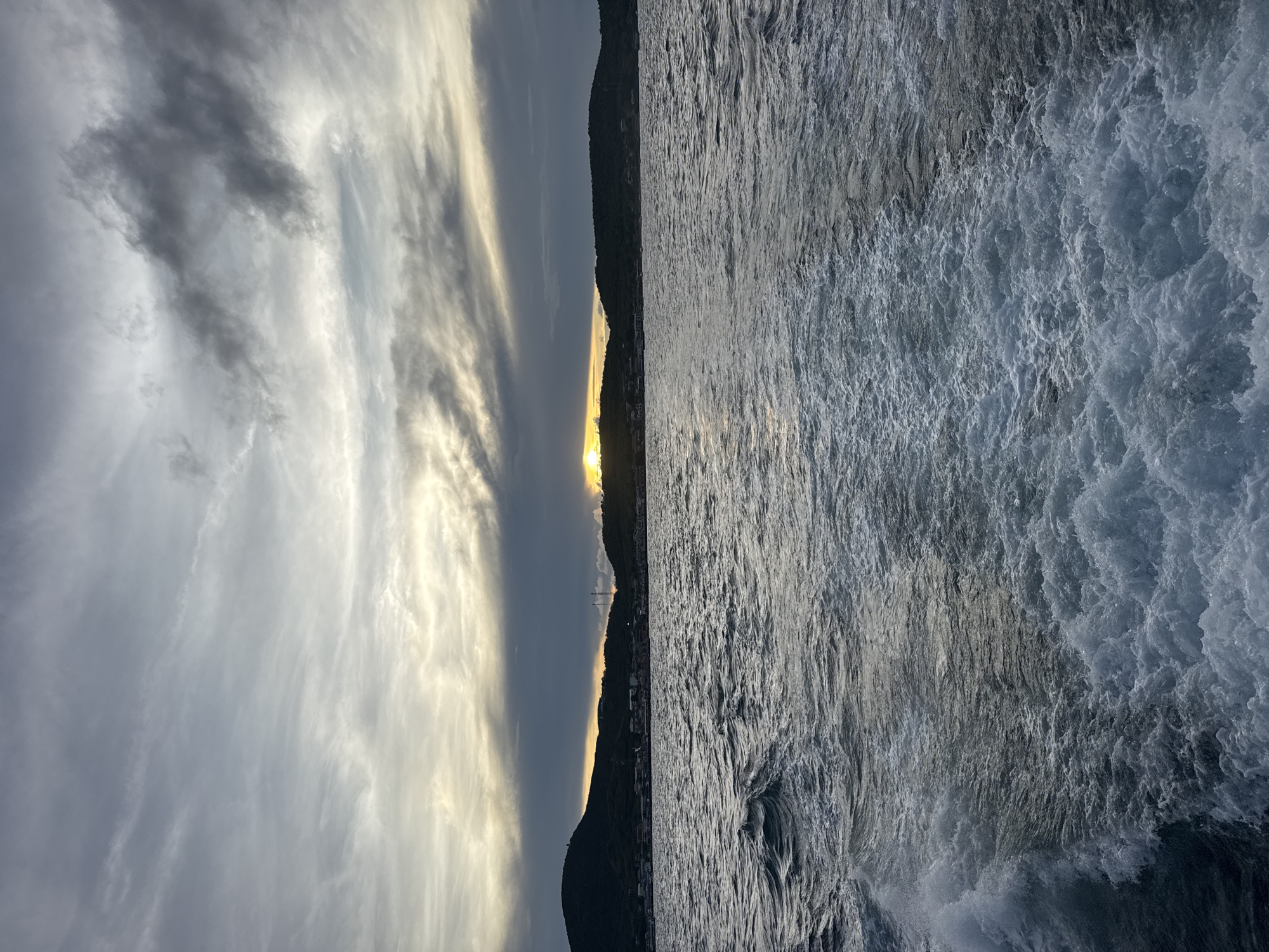 The image depicts a seascape with a dramatic sky. The horizon line is defined by a strip of land with a setting sun.
Sky: A mix of cloud formations, with light grey and white hues dominating. Some darker, grey clouds are also present.
Land: The land is dark, silhouetted against the setting sun, with hints of buildings along the coastline.
Water: The water is turbulent, with visible waves and white water spray. The color of the water ranges from dark grey to lighter shades.
Location: Given the mention of Pattaya City, Thailand, it can be assumed this is a coastal scene from that location. The image suggests a natural setting with coastal terrain, rather than a highly urbanized view.
