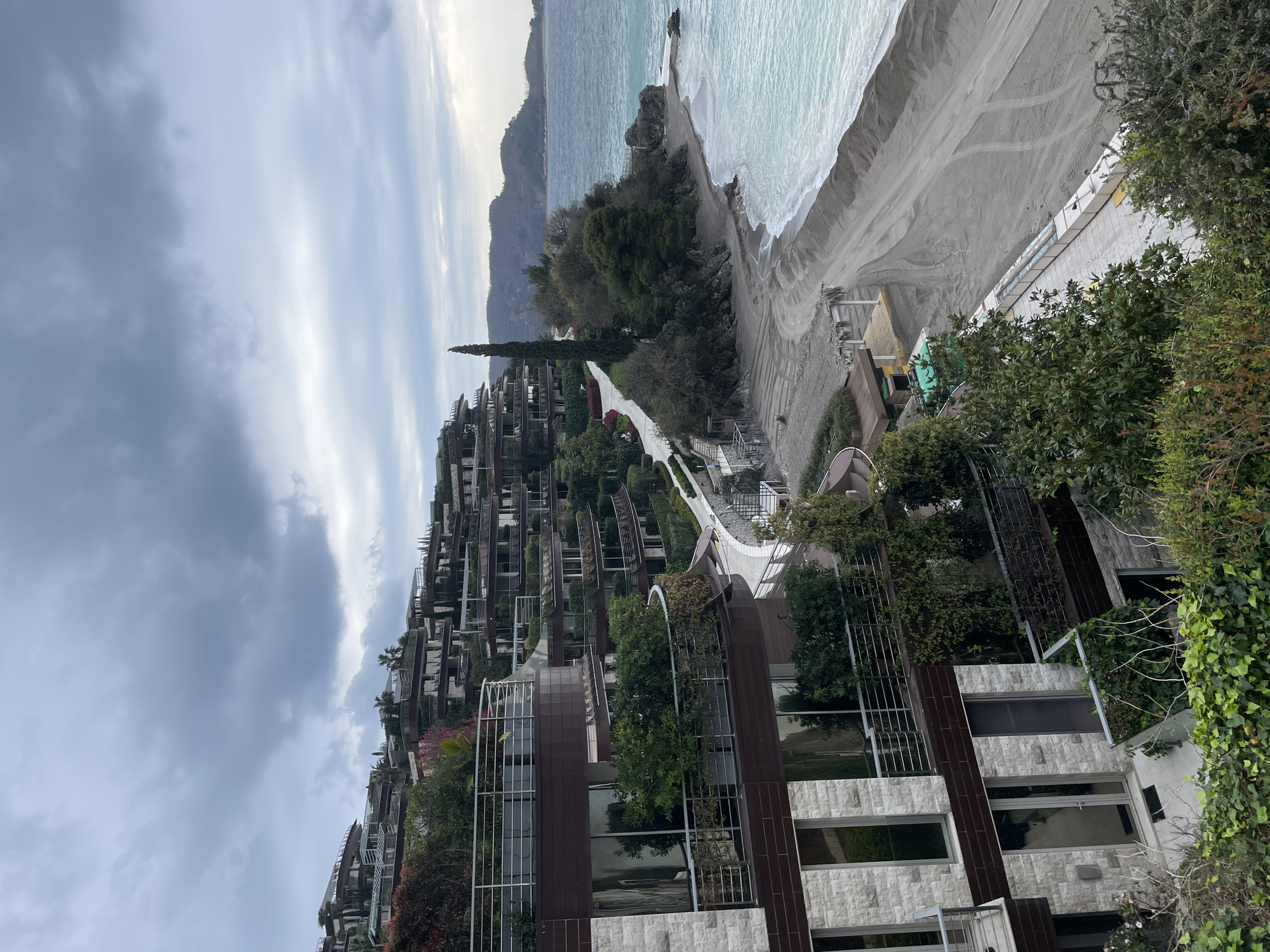 The image depicts a coastal scene in Budva, Montenegro, specifically identifying as the Dukley Gardens complex. The viewpoint is elevated, looking down towards the Adriatic Sea and along a stretch of beach.
The foreground features multi-story, terraced residential buildings constructed into a lush, green hillside. These modern structures exhibit a blend of light grey stone cladding and dark brown paneling, with numerous glass windows and balconies. The balconies are equipped with metal railings and integrate extensive green vegetation, including potted plants, bushes, and trees, giving the complex a verdant appearance. A winding, light-colored paved pathway or road snakes down the hillside, connecting the various building levels.
To the right of the buildings, the landscape transitions to a natural coastal area. A wide, grey sand and pebble beach occupies the mid-ground, showing signs of recent activity or maintenance, including visible tracks and small mounds of material. The turquoise-blue sea, characterized by light waves breaking on the shore, extends into the background. A prominent tall, slender cypress-like tree stands at the edge of the constructed area, near the beach. Further along the coastline, a rocky outcrop juts into the sea, partially defining a small cove. Distant hills or mountains are visible across the water, partially obscured by atmospheric haze.
The sky is overcast with a blanket of varied grey clouds. No individuals or active vehicles are visible anywhere in the scene. A small, partially constructed or covered structure with a thatched roof is present on the beach, along with what appears to be a green container or utility box. A white paved walkway parallels the beach in the foreground, featuring several rectangular white structures.