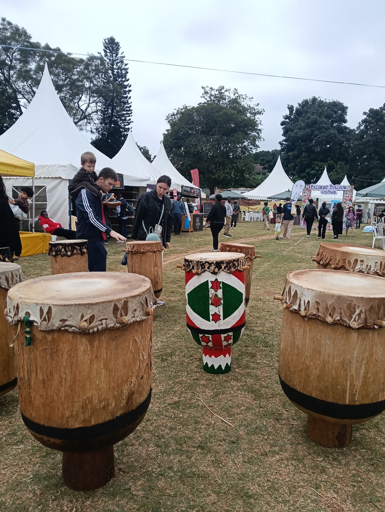 The image depicts an outdoor event, possibly a festival, taking place on a grassy area in Nairobi, Kenya. In the foreground, several large, traditional drums are prominently displayed, some standing on wooden bases and others resting on the ground. The drums appear to be made of natural materials, with stretched animal hide stretched across their tops. One drum in the center is brightly decorated with green, red, and white patterns.
In the middle ground, a group of people are interacting with the drums and the surrounding environment. A man with a young child on his shoulders is standing near a drum, appearing to be showing it to the child. A woman is standing next to him, also looking at the drums. Further back, other individuals are seen walking around, some interacting with stalls set up under white, conical tents. These tents suggest a market or festival setup. A banner in the background reads "BIZARRE BAZAAR FESTIVAL," confirming the nature of the event.
The background features lush green trees and a cloudy sky, characteristic of Nairobi's climate. The overall atmosphere seems casual and festive, with people enjoying the outdoors and the cultural displays. The presence of tents and various people suggests a public gathering or market event.