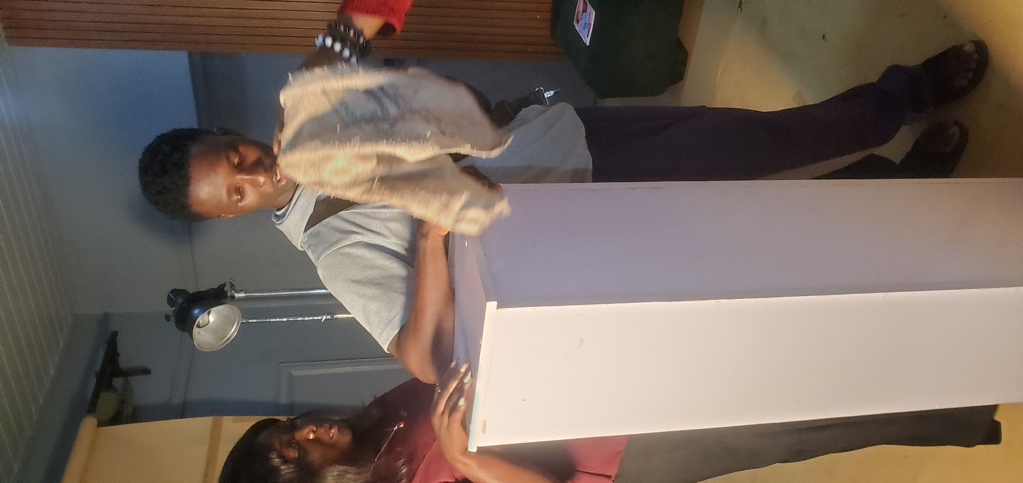 Two individuals, an adult female and a young adult male, are standing in what appears to be a studio or indoor setting in Bukuru, Nigeria. The female, on the left, has dark hair and is wearing a maroon shirt and black pants. She is leaning on a tall, pale pink rectangular object, with her left hand resting on its surface. The young male, on the right, has short dark hair and is wearing a light blue hooded shirt and dark pants. He is holding a tan, roughly textured piece of cloth in both hands, which he appears to be either examining or cleaning. His posture suggests he is either in the process of a task or demonstrating something. Behind them, a studio lamp on a stand is visible, along with a portion of a wall with vertical wooden paneling. The floor is a light yellow color. The overall environment suggests a preparation or work in progress.
