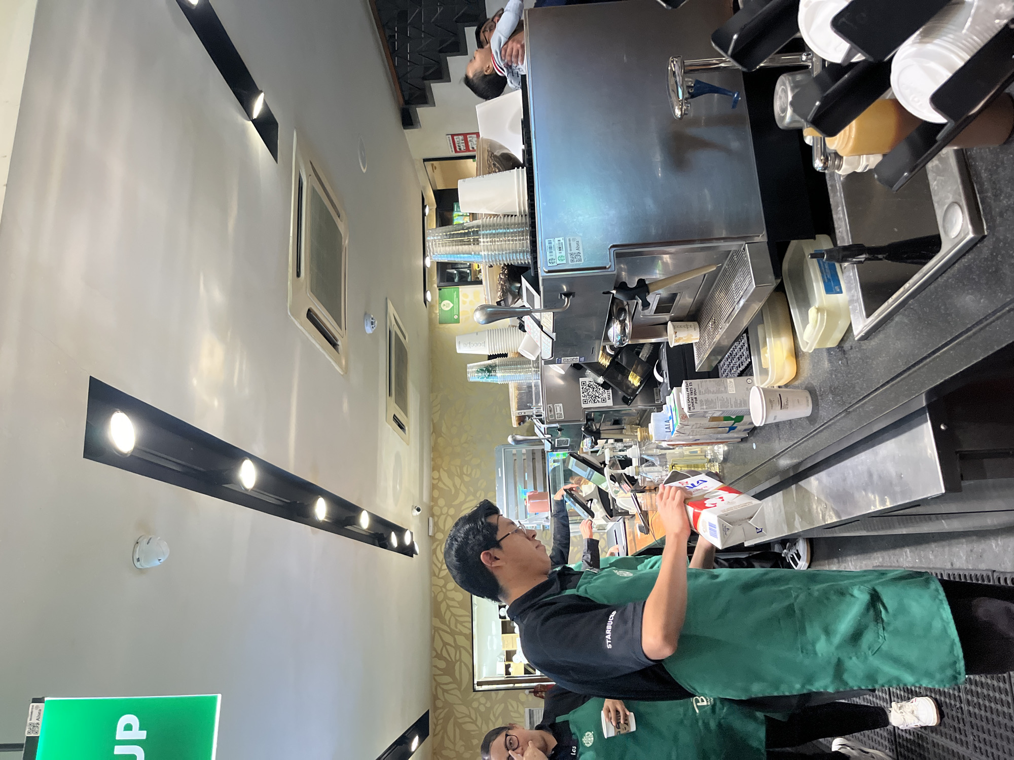 Here is a description of the image:
**Setting:** A Starbucks store, potentially located in Puebla, Mexico, as the milk carton reads “Alsea.”
**People:** Two Starbucks employees wearing green aprons and black shirts. One is holding a white and red milk carton, presumably preparing a beverage. The second barista is partially visible in the lower portion of the image.
**Objects:** Starbucks branded items, including paper cups, milk cartons, an espresso machine, and various containers for syrups and sauces. There is also a "PICK UP" sign partially visible.
**Actions:** One barista is about to pour milk from the carton.
**Environment:** The store has modern lighting fixtures. An air conditioning unit is visible on the wall.