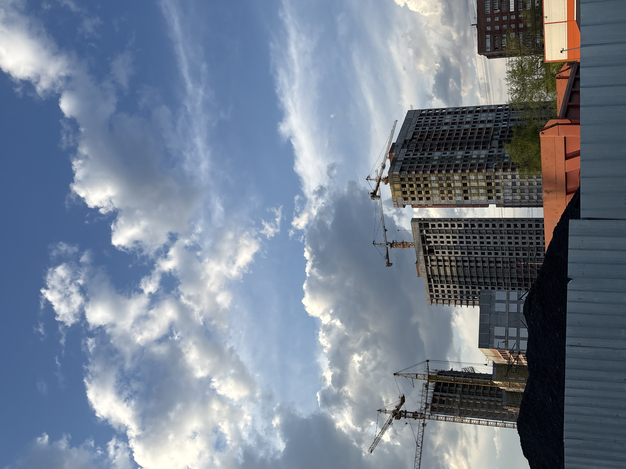 The image shows a construction site in Perm, Russia. Three unfinished high-rise buildings are visible, each accompanied by construction cranes. The buildings are in various stages of completion, with exposed concrete frames. In the foreground, construction materials and a metal fence are present. An older residential building can be seen in the background. The sky is partly cloudy with blue patches visible.