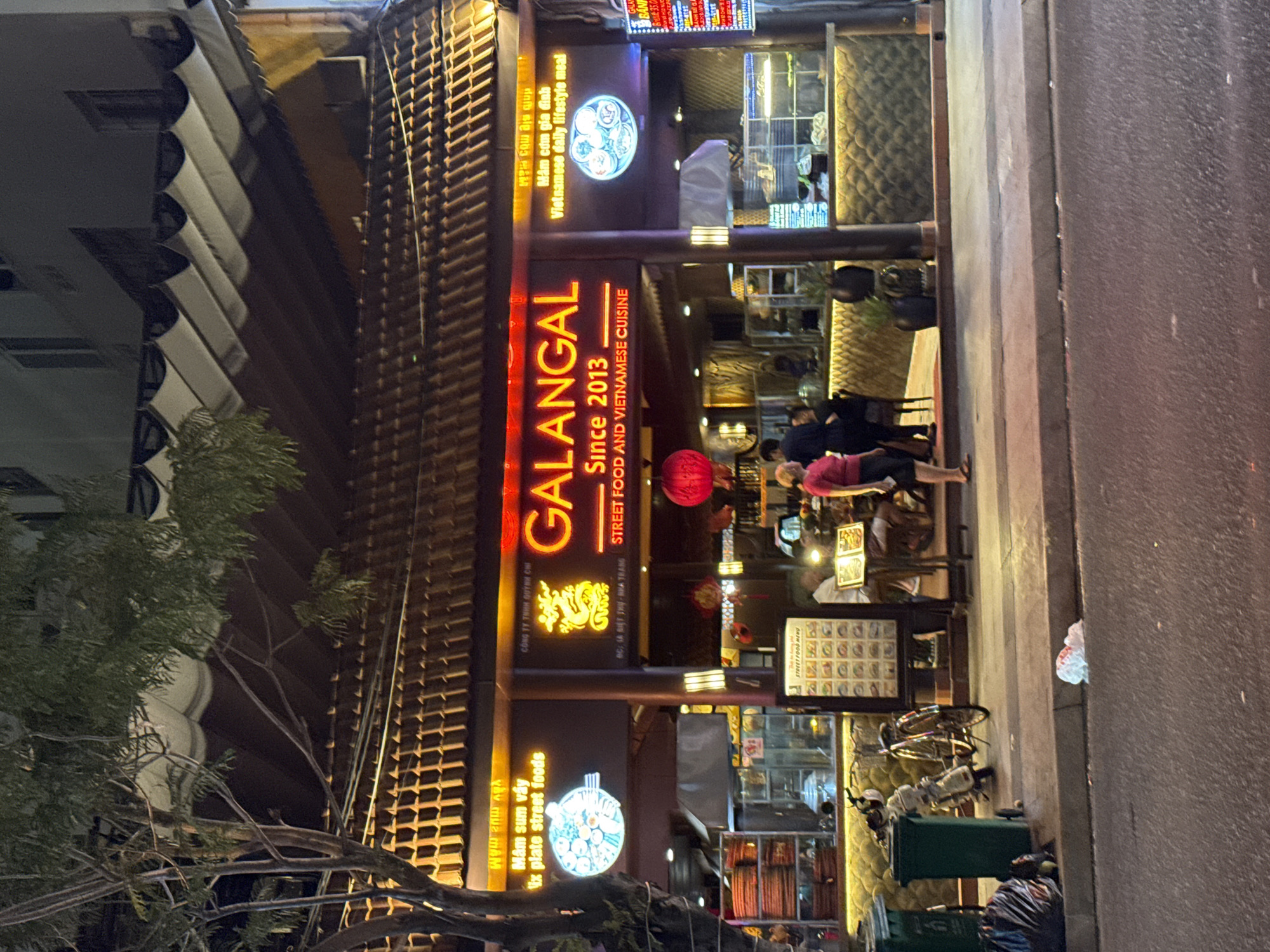 The image shows the front of the Galangal restaurant in Nha Trang Ward, Vietnam. The restaurant's sign, illuminated in neon orange, reads "GALANGAL Since 2013 STREET FOOD AND VIETNAMESE CUISINE." People are seated at tables inside the restaurant. A sign to the left of the main sign advertises "Mâm sum vầy ix plate street foods." Bicycles and trash bins are near the sidewalk to the right of the restaurant entrance. A tree is on the left side of the image. The overall impression is of an evening street scene.