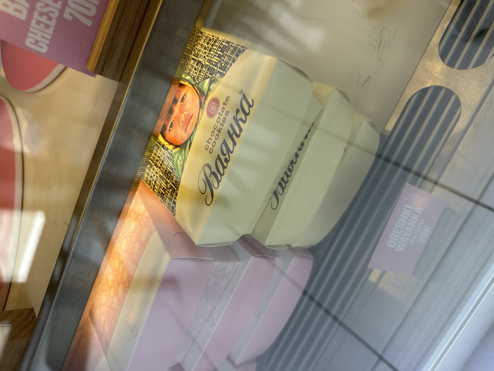A view of a display case, likely inside a shop in Mengwi, Indonesia, showcasing packages of “Валенка” chocolate cookies. The cookies are packaged in yellow and white boxes and feature an image of a child’s face. Adjacent to the cookies are pink boxes, possibly containing other baked goods. A price tag is visible, indicating “ORESHKI WAYANKA chocolate 70K.” The display case is clean but has some reflection on its glass surface.