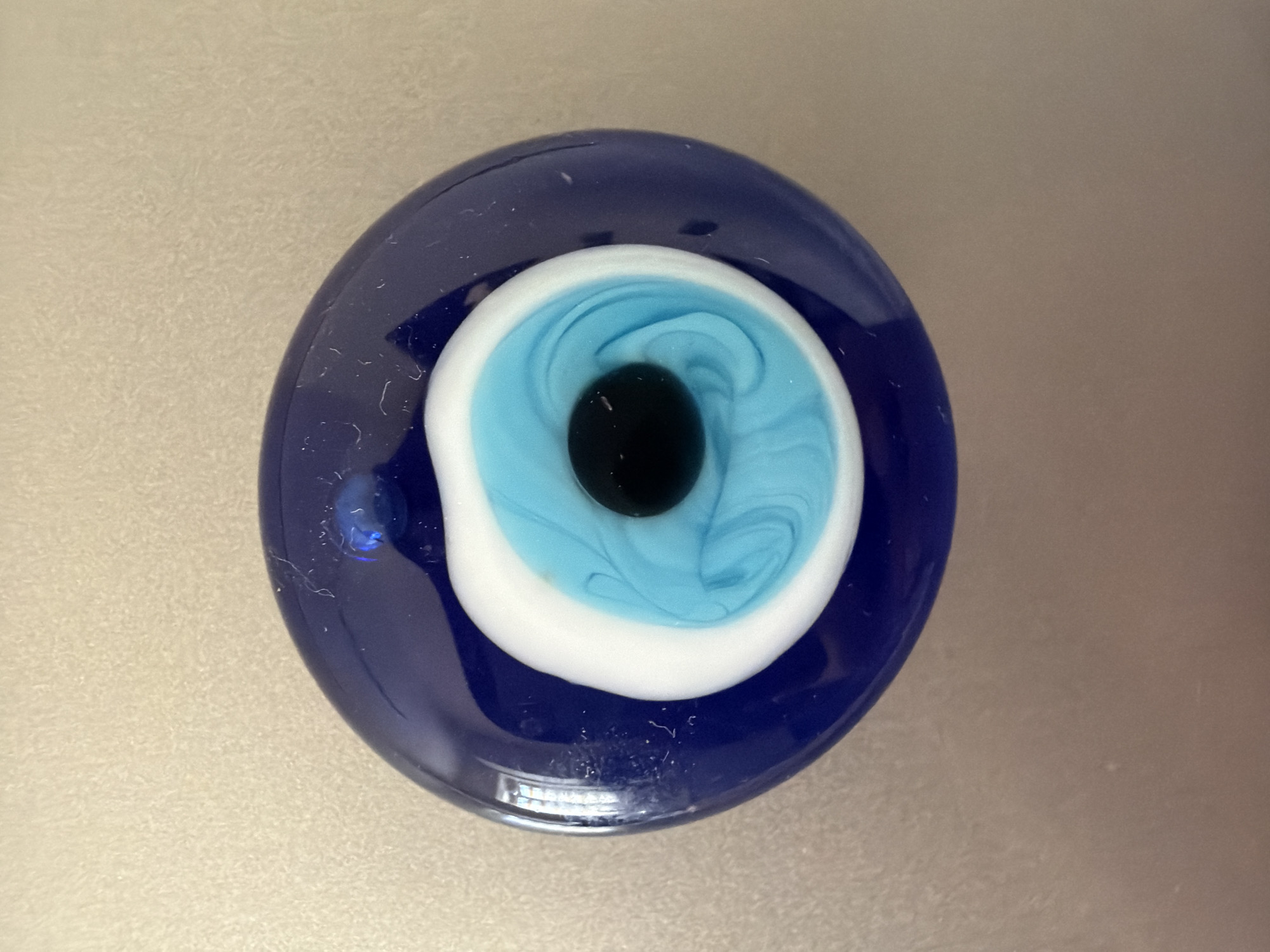 A close-up overhead shot shows a circular blue glass evil eye charm resting on a textured, beige surface. The charm is dark blue and features concentric rings of white, bright turquoise, and a central black pupil, designed to resemble an eye. A small, clear blue bead is visible at the top of the charm. The background surface appears to be a neutral, possibly metallic or stone material, with a subtle texture. The lighting is bright, creating reflections on the glossy surface of the charm. The location in Pazardzhik, Bulgaria, is not discernible from the image itself.