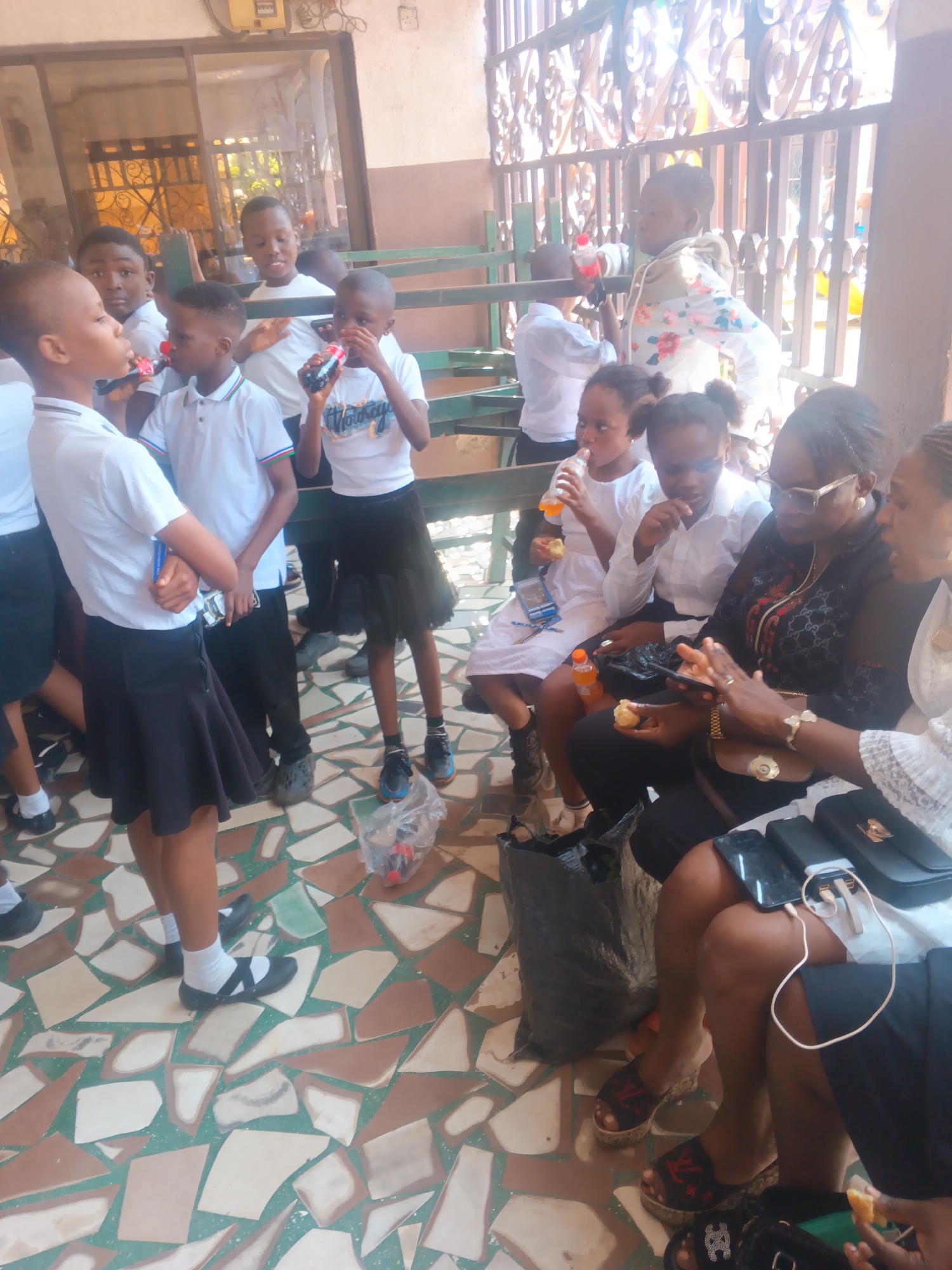 The image depicts a group of children and adults gathered outdoors, possibly in a schoolyard or a public space in Onitsha, Nigeria. The ground is paved with broken tiles in various shades of brown, cream, and green. In the background, a metal fence with ornate patterns suggests a structured environment. Several children, dressed in white tops and black bottoms (skirts and trousers), are holding and drinking from bottles, likely soft drinks. Some appear to be interacting with each other, while others are focused on their drinks. Adults are also present, seated and observing the children. One woman in the foreground is looking at a smartphone. The lighting suggests it is daytime.
