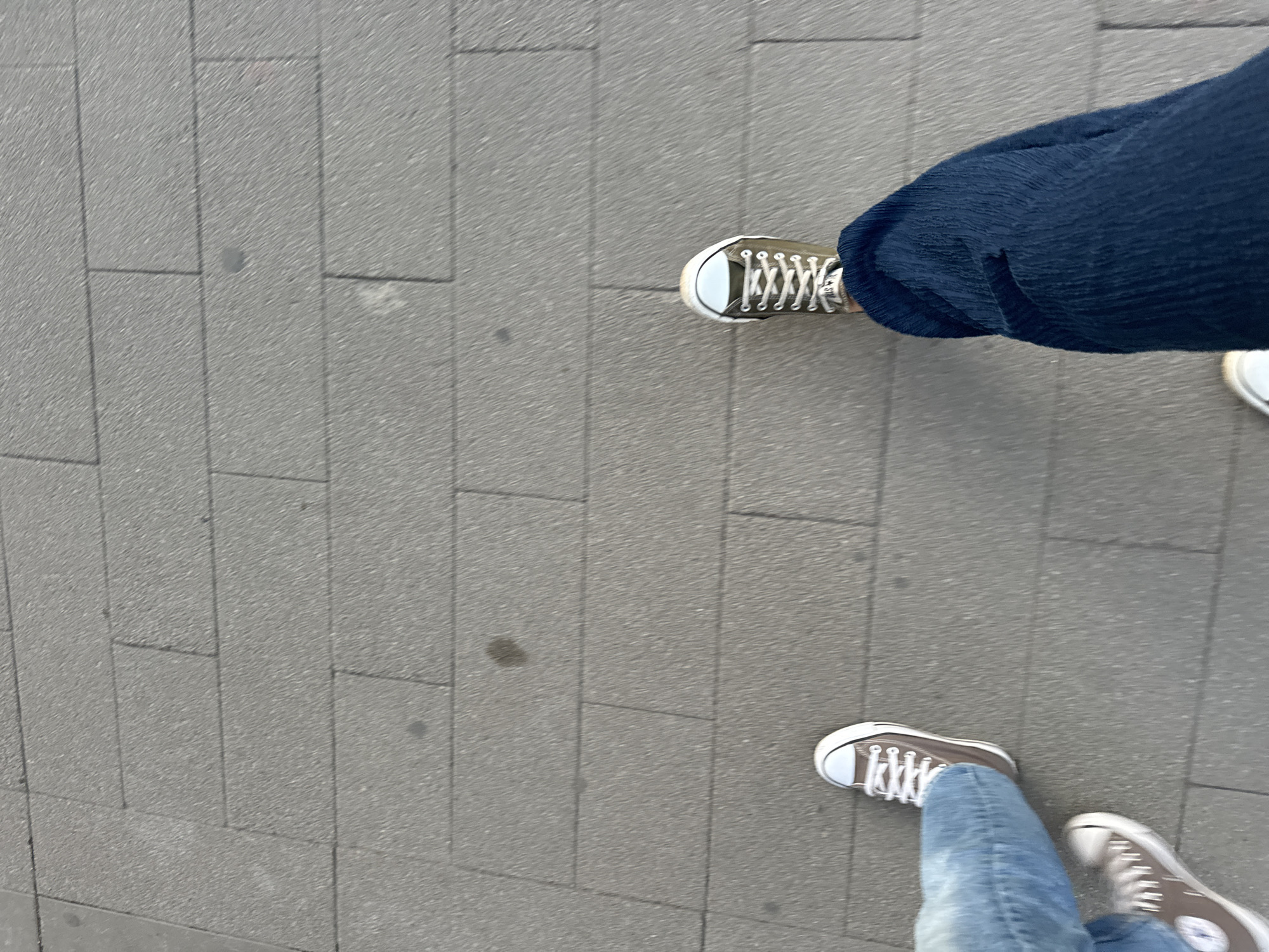 Two pairs of feet are visible, walking on a grey paved sidewalk. The person on the left is wearing blue jeans and brown low-top canvas sneakers. The person on the right is wearing dark blue pants and olive green low-top canvas sneakers. The image appears to be taken from a first-person perspective, looking down at the feet and the ground. The environment suggests an outdoor urban setting.