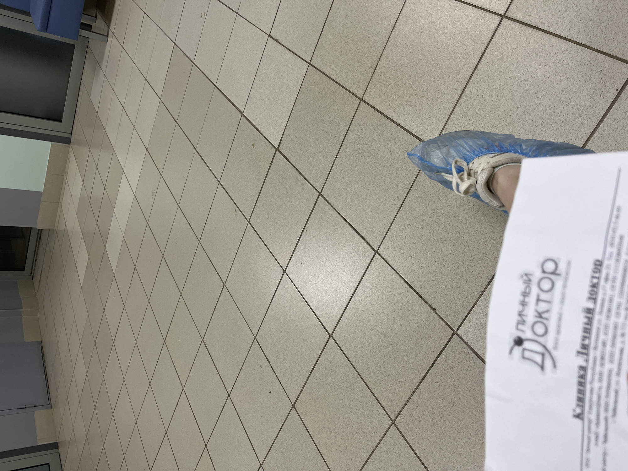 The image shows a person wearing a blue shoe cover on their right foot while standing on a tiled floor, likely in a medical clinic. They are holding a document from "Личный Доктор" (Personal Doctor), a clinic located in the city of Chaikovsky, Russia. The floor tiles are light grey, and a waiting area with blue chairs is visible in the background.
