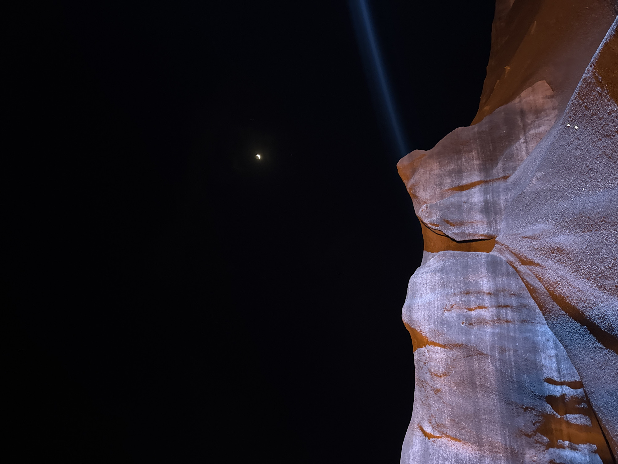 At night in Teguise, Spain, a spotlight shines down upon the monument El Monumento al Campesino. The monument, composed of rough, textured concrete, is partially lit with an orange hue. In the dark night sky, a faint crescent moon is visible near a small dot. The background is entirely dark.