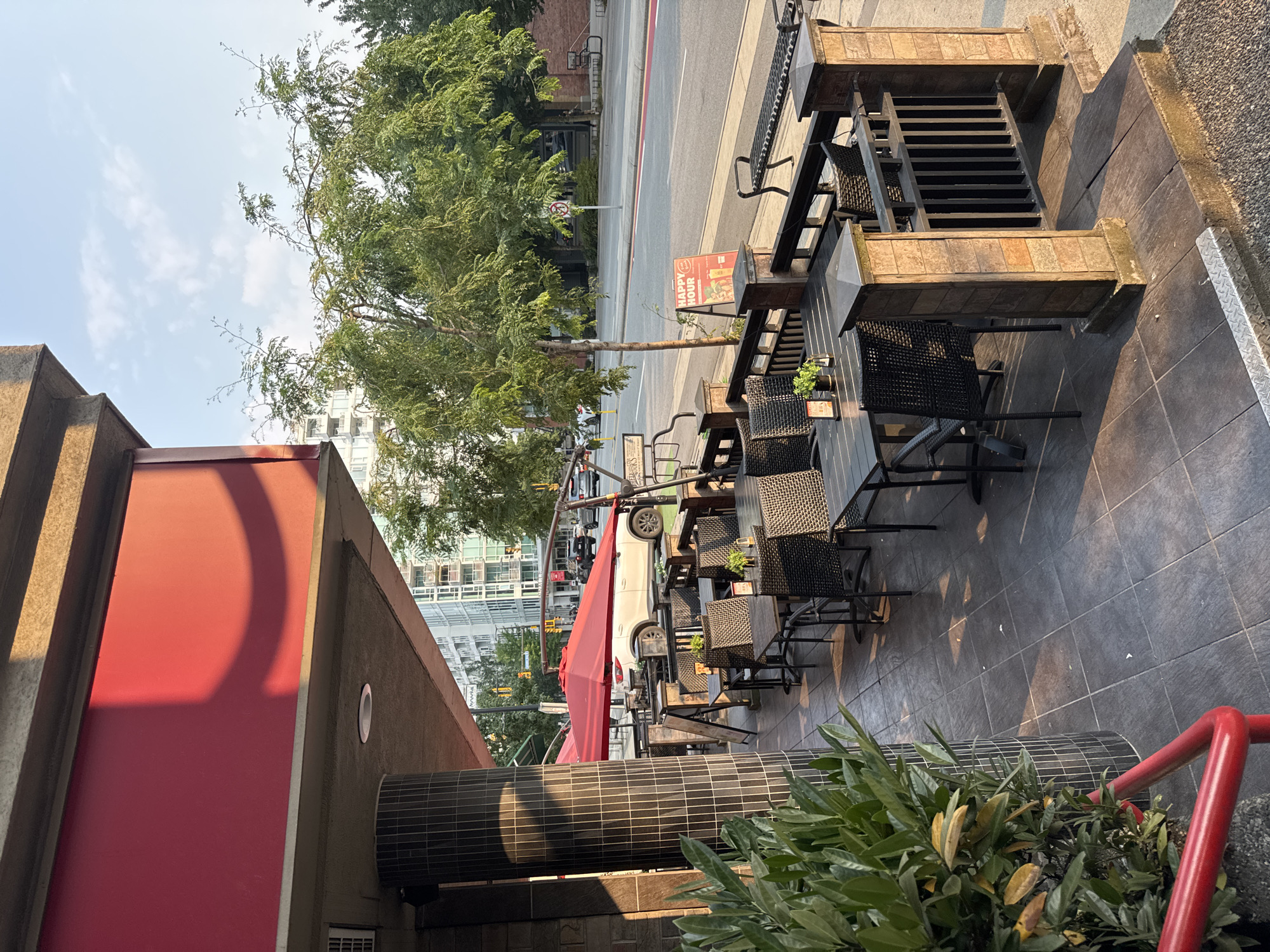 An exterior shot captures the patio seating area of a restaurant on a sunny day, with urban buildings and trees visible in the background. The patio is furnished with several dark-colored tables and chairs, some set with small potted plants and menus. A large red umbrella provides shade over some of the tables. A "Happy Hour" sign is placed on an easel near the edge of the patio. The patio is paved with dark tiles and features stone pillars and a black metal railing. In the background, a white SUV is parked in front of the restaurant. The street in front of the restaurant has a red painted lane and traffic signals. The location is the city of North Vancouver, Canada, likely on a commercial street with both residential and commercial buildings.