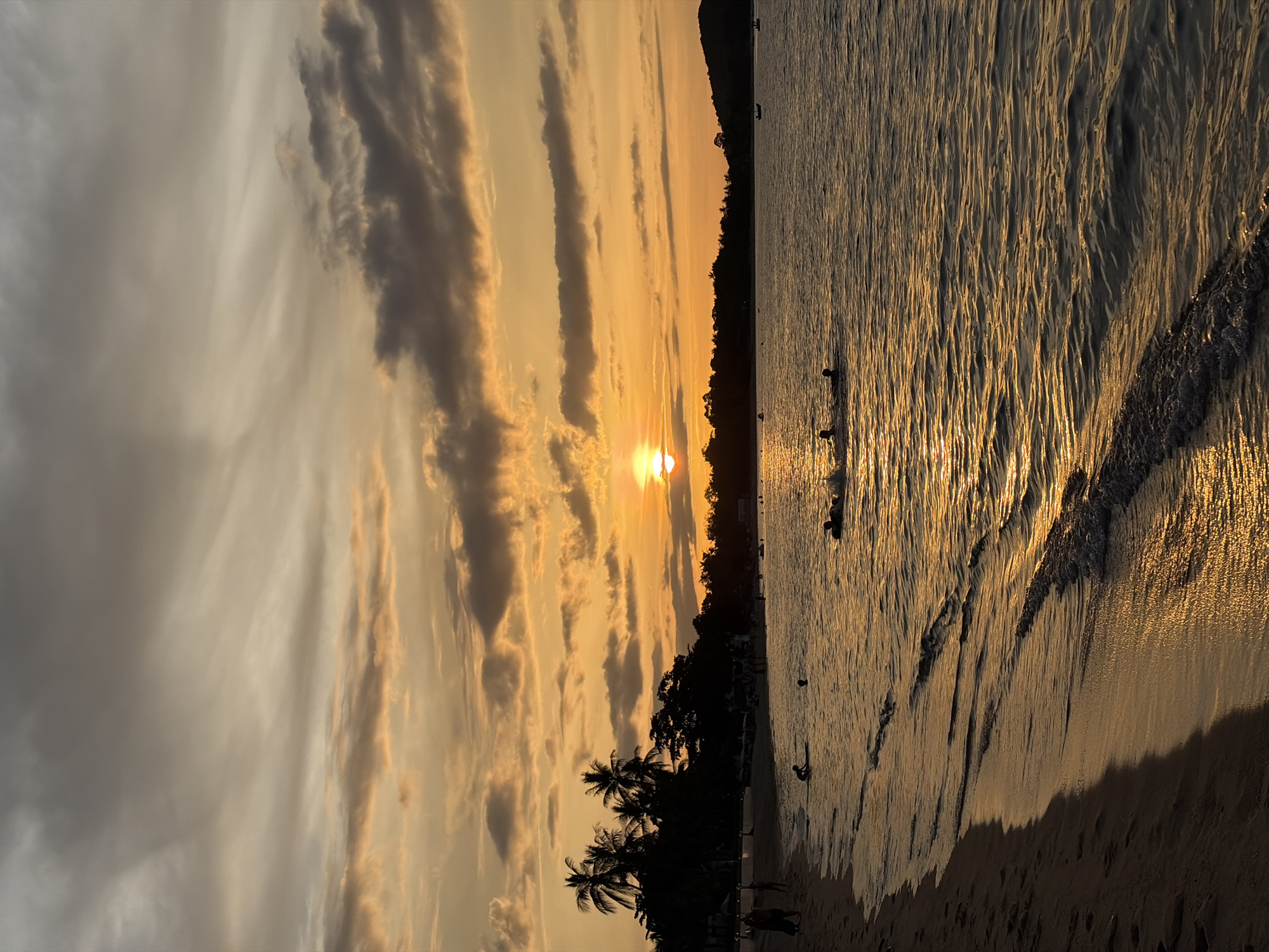 This image captures a sunset scene at a beach in Ko Samui District, Thailand. The sky is filled with dramatic clouds painted in shades of orange, yellow, and gray, with the sun partially obscured by clouds, casting a warm glow. The ocean is calm, reflecting the colors of the sunset, with gentle waves lapping at the shore.
On the beach, several people are visible, some walking along the sand and others swimming or playing in the water. Palm trees and other vegetation form a silhouetted treeline along the coast. In the distance, a few boats are moored in the water. The overall impression is one of a tranquil evening at a tropical beach.