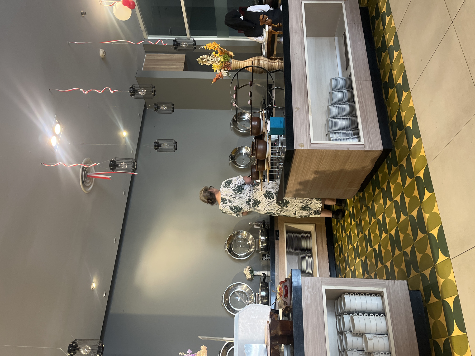 A woman in a floral dress is serving herself from a buffet in a restaurant in Gapurosukolilo, Indonesia. The buffet has several chafing dishes, pots, and serving utensils. There are stacks of plates and cups on shelves below the counter. The room is decorated with hanging lanterns and red and white streamers. The floor has a geometric pattern of green and gold tiles.