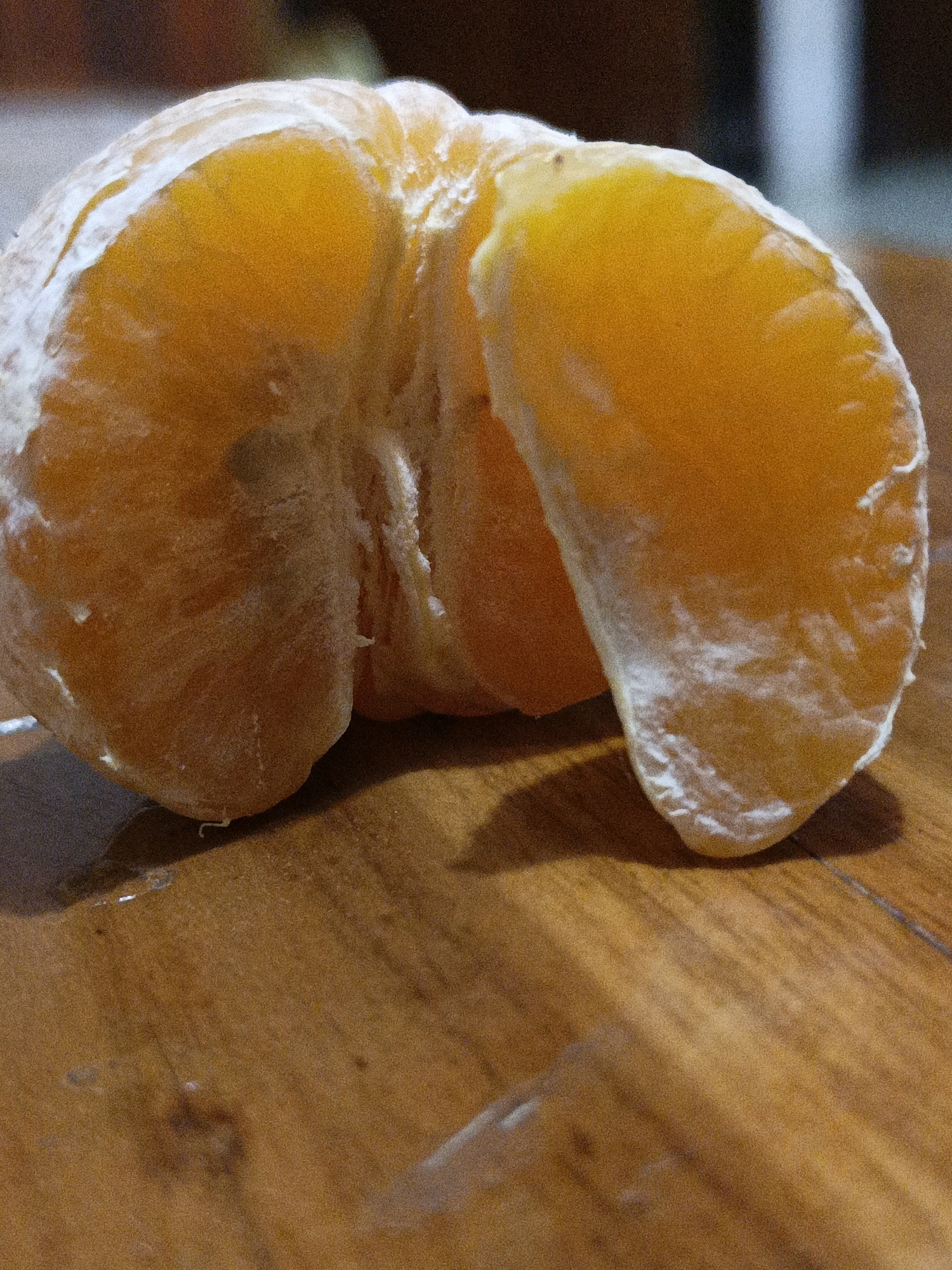 A peeled tangerine is positioned on a wooden surface. The tangerine is partially opened, revealing its segments. The surface appears to be a table or countertop. The lighting suggests an indoor setting, possibly a kitchen or dining area. The image's origin is not specified and therefore cannot be linked to Malang, Indonesia.