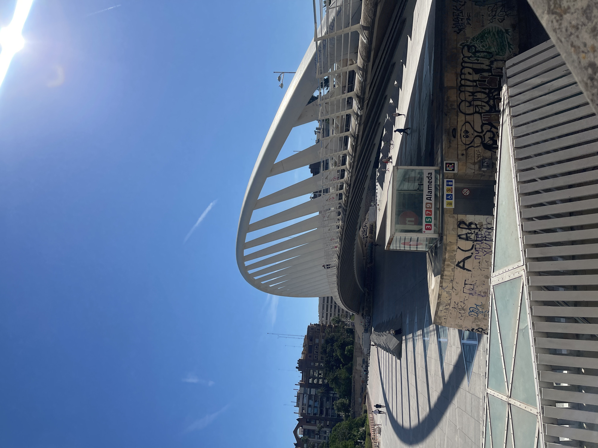 The image shows a view of the Pont de l'Exposició in València, Spain. The modern white bridge with its arching ribs is the prominent feature. Below the bridge is the "Alameda" metro station entrance, indicated by signage with line numbers 3, 5, 7, and 9. Graffiti is visible on the walls near the metro entrance. Buildings and trees are visible in the background along the Turia Gardens. Two pedestrians are walking along the sidewalk. The sky is blue. The sun's glare is visible in the upper left corner.