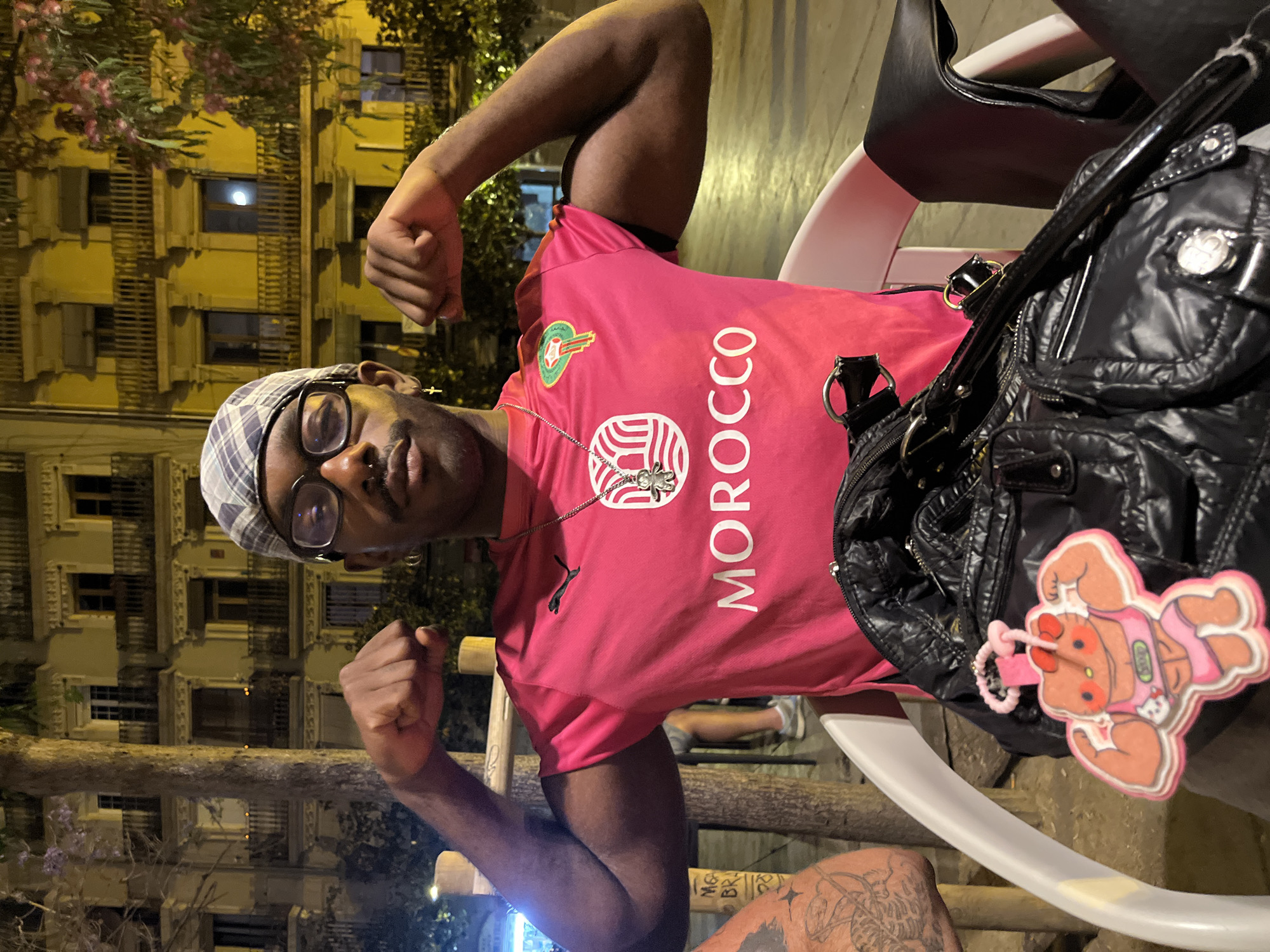 A man wearing a pink Puma T-shirt with "MOROCCO" printed in white sits in a white chair, flexing his fists. He has a plaid hat, glasses, and a cross necklace. A black purse with a pink Hello Kitty weightlifter keychain is next to him. The background shows a multi-story building typical of Barcelona architecture, illuminated at night. He is likely at an outdoor cafe.