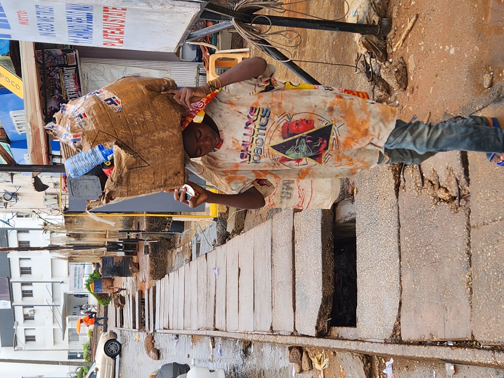 A person carries a large sack on their head in an urban setting. The individual is wearing a graphic t-shirt with the words "SHLUXE ROBOTICS" and a print featuring human faces, and what appear to be blue jeans and sandals. The sack is made of cardboard and plastic, and has a blue water bottle visible. The person appears to be walking along a street next to a sidewalk or gutter. There's a drain or opening in the sidewalk. In the background, there are buildings and signs, including one that reads "PLATEAUSTATE" and "IN GOD WE TRUST." There are also cars and other people in the background. The ground appears wet.