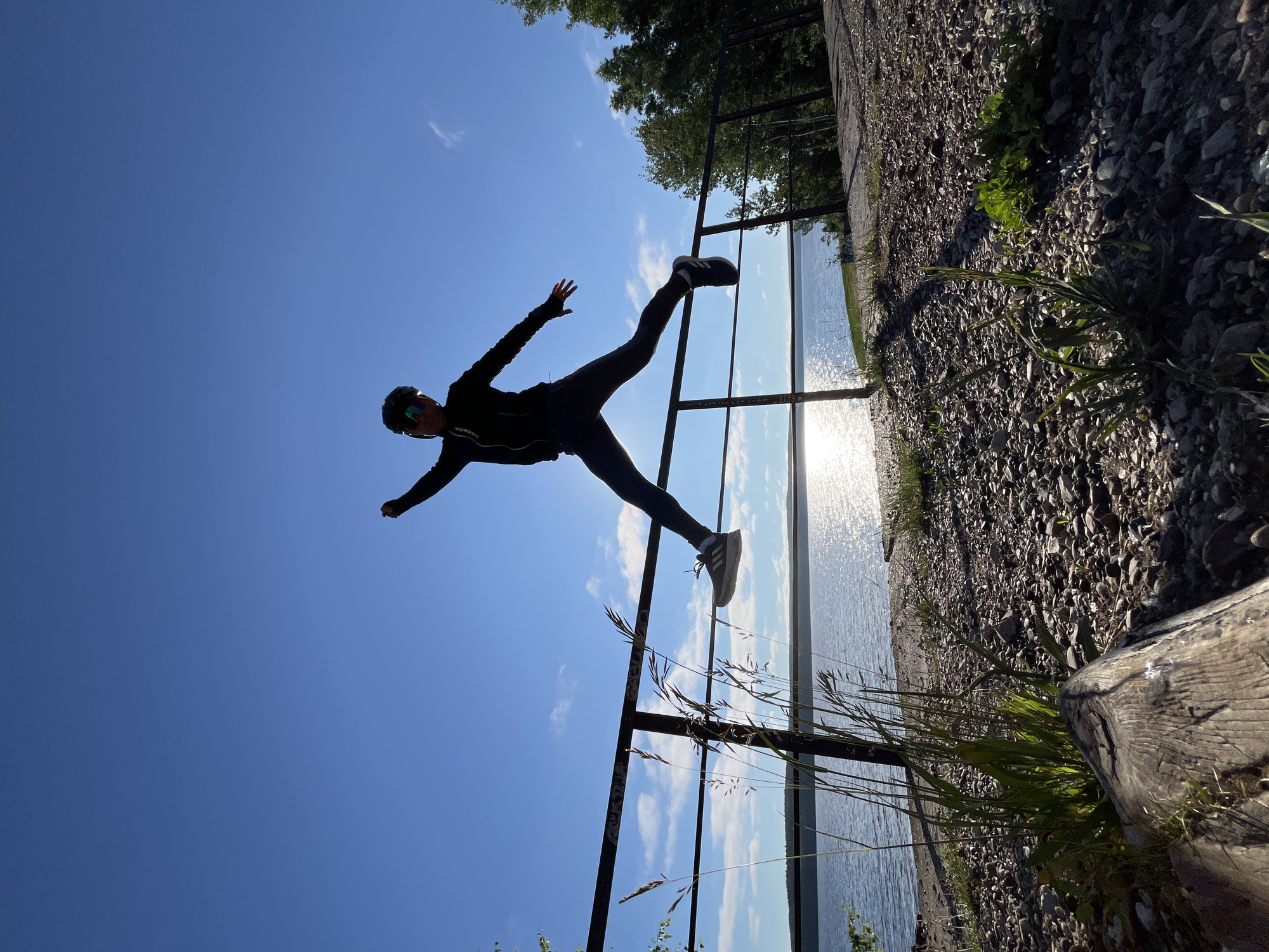 A person in silhouette is captured mid-jump over a railing, against a clear blue sky. The person is wearing dark clothing, a helmet, and sunglasses, with one leg extended forward and the other bent. The railing separates a gravel path with some vegetation from a body of water, likely the Kama River in Chaikovsky, Russia, as indicated by the sunlight reflecting on its surface. Trees are visible in the background.