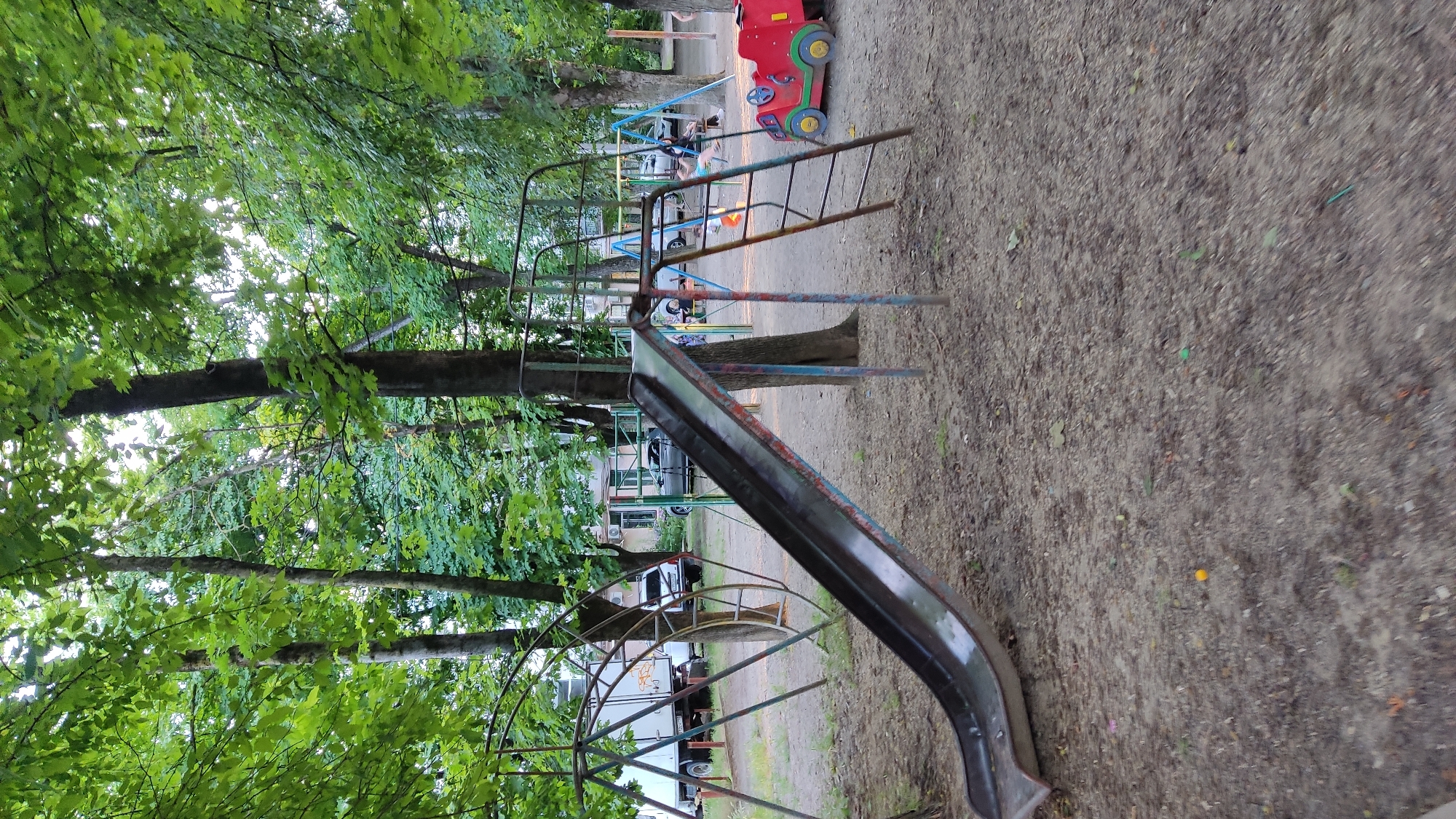 A playground scene in Krasnodar, Russia, depicts a weathered metal slide as the central focus. The slide, showing signs of rust and faded paint, descends towards a sandy ground covered with dirt and scattered leaves. To the left of the slide, a triangular climbing frame with metal bars is visible, and behind it, a white truck with graffiti is parked. In the background, several cars are parked, and apartment buildings line the perimeter. Further to the right, a swing set with two visible swings and a red toy car are present. The playground is surrounded by mature trees with dense green foliage, casting shadows and creating a dappled lighting effect. The sky is overcast, suggesting a cloudy day. One person is seen on the swing set.