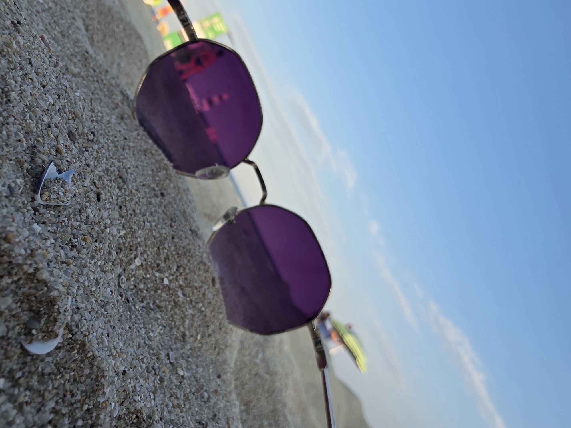 A low-angle, close-up shot focuses on a pair of sunglasses with purple lenses resting on coarse, light-colored sand. The sunglasses are positioned in the foreground, with their lenses reflecting a bright, hazy scene. In the reflection of the left lens, a pinkish figure and a structure resembling a lighthouse can be discerned, along with some blurry green and yellow shapes that might be beach umbrellas or structures. The background shows a clear, pale blue sky with faint wispy clouds and a suggestion of the horizon. The environment appears to be a beach. It is not possible to determine the specific location within Obrochishte, Bulgaria from the provided image.