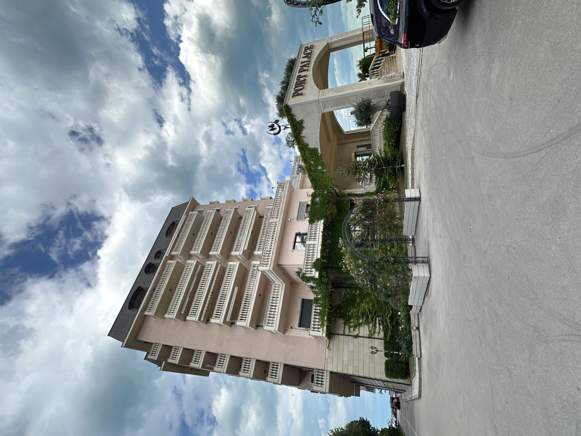 The image shows the "Port Palace" hotel in Varna, Bulgaria, as indicated by the building's signage. The hotel is a light pink building with multiple balconies, situated next to a road. A black car is parked near the hotel entrance. The sky has a mix of blue patches and heavy, overcast clouds. The hotel facade is adorned with ivy, and a decorative metal structure with flowering vines is also visible. The hotel entrance features an arched entryway with the hotel name displayed prominently. The overall scene appears to be a sunny day with a mix of clouds, capturing the architectural details of the hotel and the immediate surroundings.