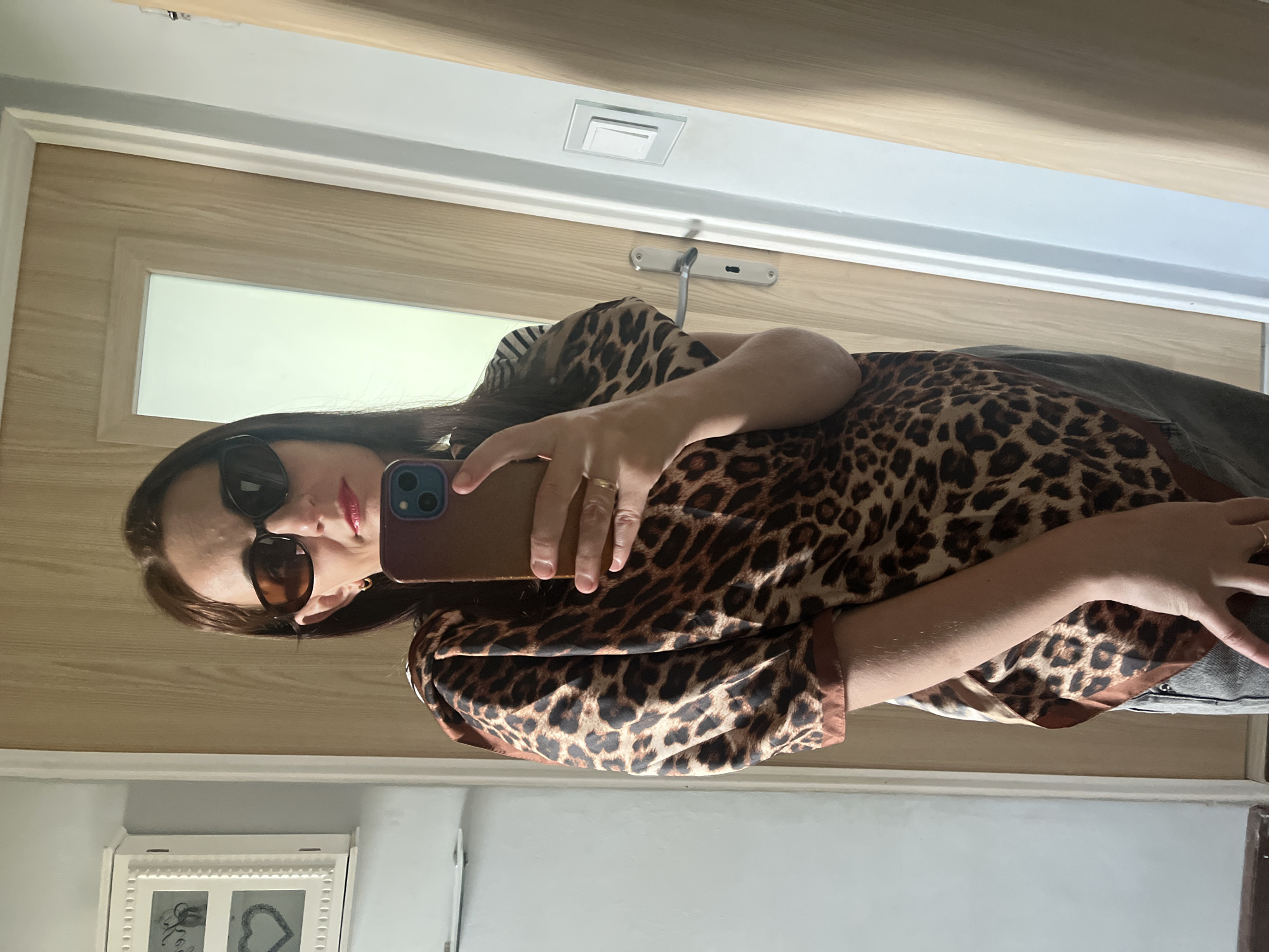 A mirror selfie captures a woman indoors, standing upright and looking into the camera held in her right hand. She has dark hair, wears dark sunglasses, and has red lipstick on. Her attire consists of a leopard print long-sleeved top or dress, paired with dark-colored trousers or a skirt, partially visible. On her right ring finger, she wears a gold-toned ring. She holds a smartphone with a reddish-brown or gold case and a blue camera module.
The background, visible in the mirror's reflection, shows a light-colored wooden door with a rectangular frosted glass panel. Above the door frame, a square white light switch is mounted on a white wall. A silver handle or latch mechanism is visible on the door. In the lower left corner of the reflection, a white decorative picture frame is seen, containing what appears to be a heart shape and another design. The walls in the reflection are primarily white above the door frame and a light gray below. The overall setting appears to be a casual indoor environment, likely a home. There is no visible text.