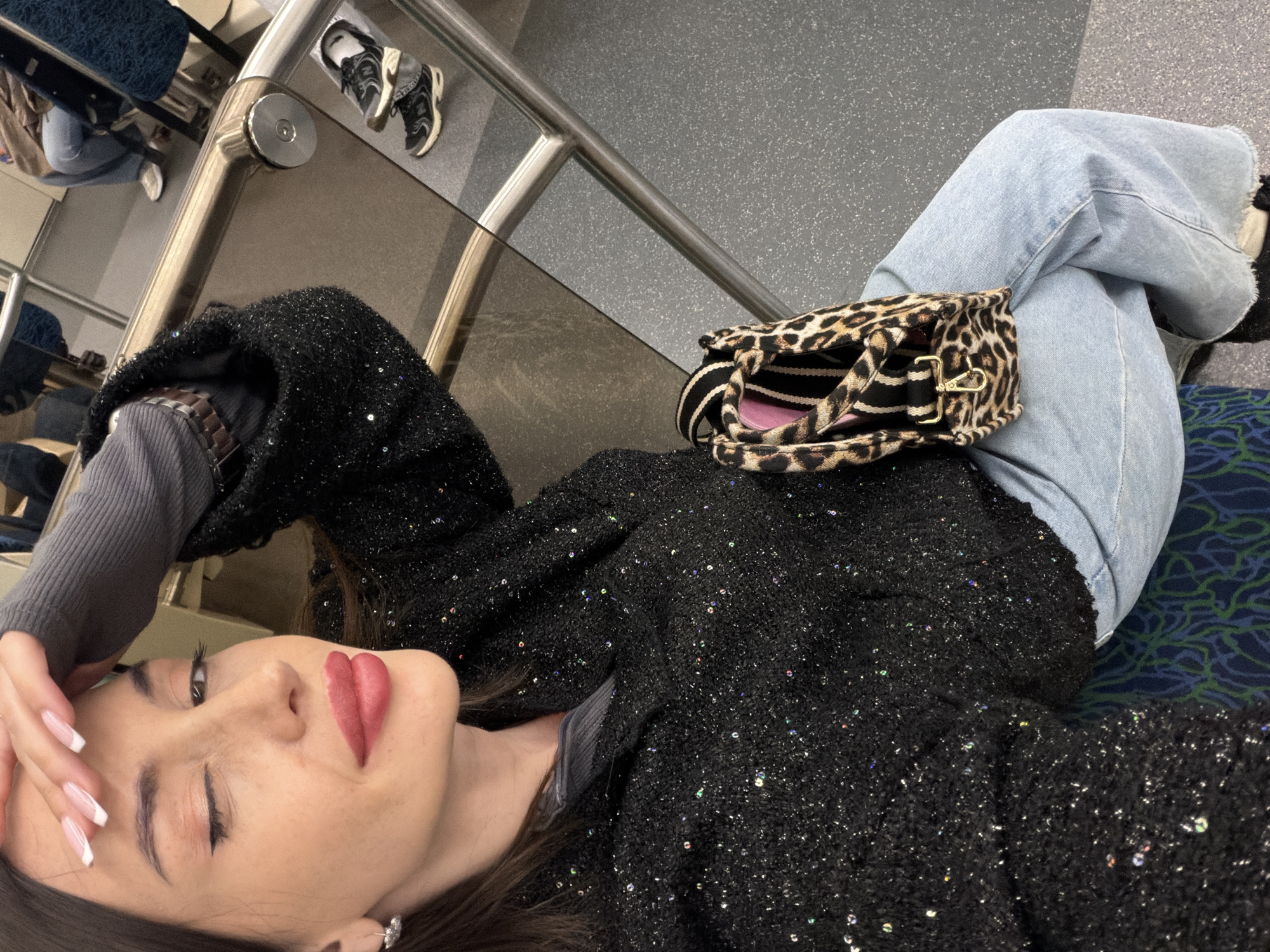 A woman lies on a seat in what appears to be a public transportation vehicle. She is positioned on her side, with her head resting on her hand. She wears a black sequined jacket over a gray long-sleeved shirt and light blue jeans. A leopard-print handbag rests on her legs. The interior of the vehicle is visible, including metallic handrails and seats with patterned upholstery. In the background, shoes are placed on a platform near the railing. The location is likely a public transportation vehicle in Saint Petersburg, Russia, based on the interior design of the vehicle.
