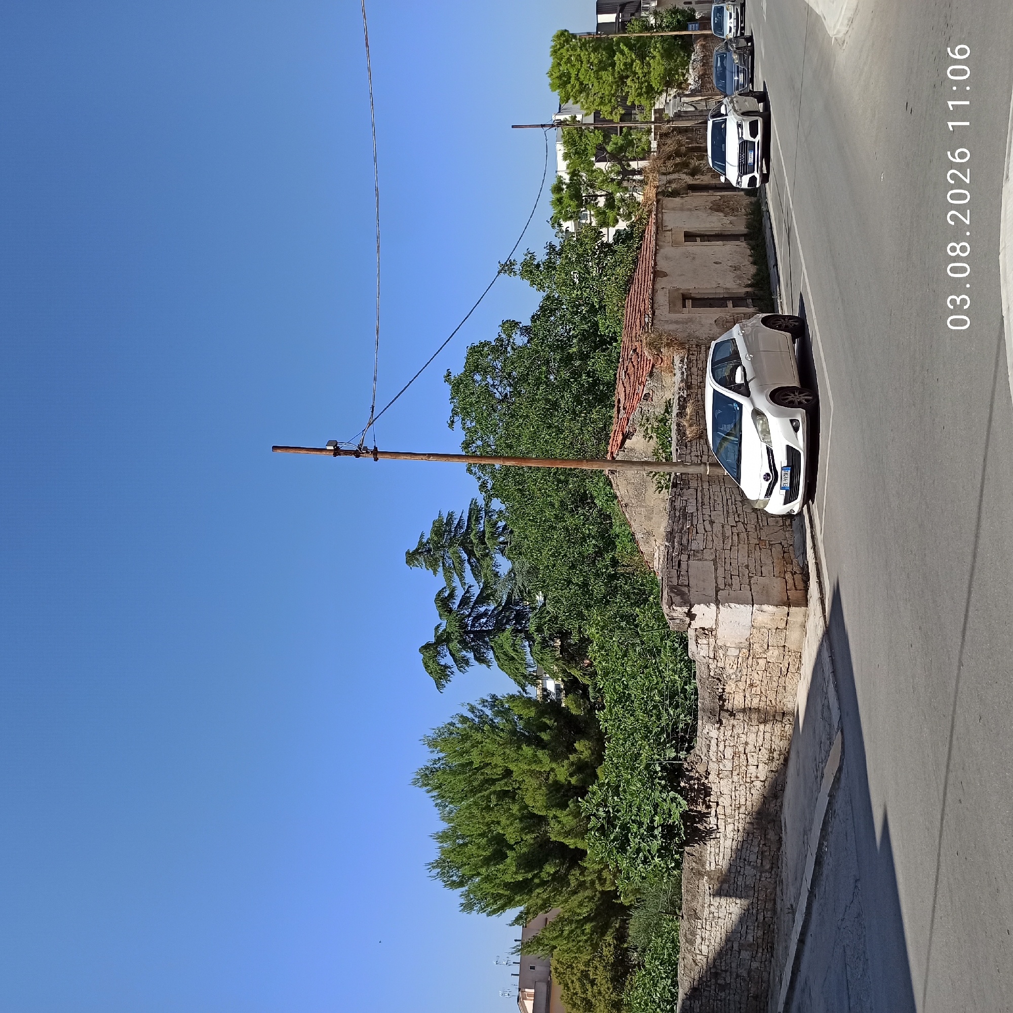 The image captures a street scene in Ruvo di Puglia, Italy, on August 3, 2026, at 11:06 AM. The sky is clear and blue, indicating fair weather. In the foreground, a paved road curves to the right, with a light gray curb and sidewalk. Parked on the right side of the road, within marked parking spaces, is a white Toyota Yaris. Further down the road, other vehicles are visible, including a white Audi, a dark gray sedan, and a yellow hatchback.
Along the left side of the road, a low, weathered stone wall borders a property. Behind the wall, a dense arrangement of green trees and foliage creates a natural barrier. Among the trees, an old, dilapidated building with a red tiled roof is partially visible. A wooden utility pole with some wires attached stands near the building and the stone wall. The background consists of more trees and modern buildings. The overall impression is a sunny day in a town with a mix of old and new architecture, and active street life with vehicles present.