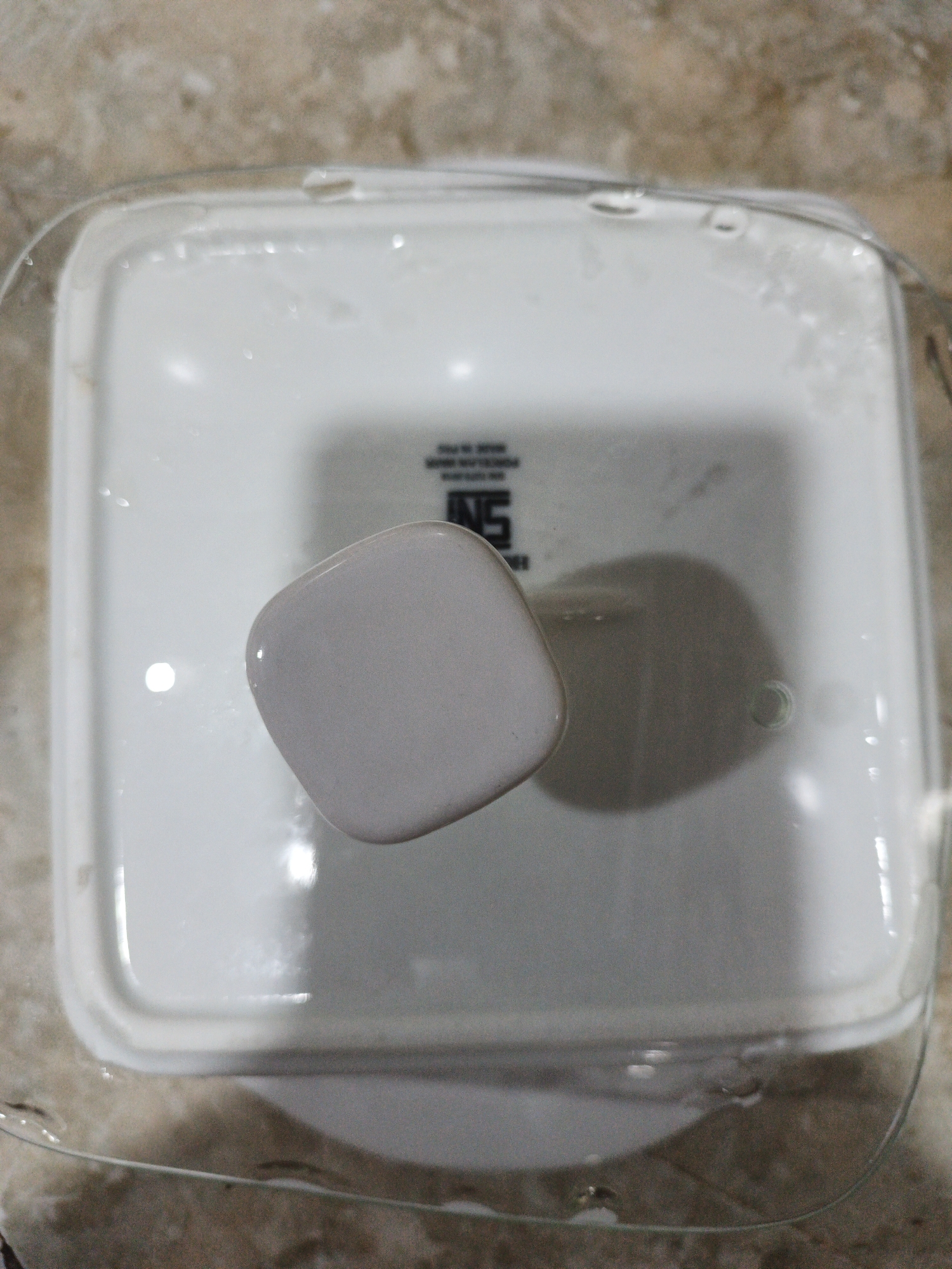 The image shows the lid of a white container, possibly a food storage box, placed on a marble-patterned surface. A clear plastic rim or edge is visible around the lid, and there are water droplets on both the lid and the surface. A small, rounded, light gray object is resting on the lid, casting a shadow. The lid has a faint logo with the letters "SN" and other text that is difficult to read. There are no people or active interactions depicted in the image. The scene appears to be indoors, given the lighting and indoor surface. The location in Kelapapati, Indonesia, cannot be determined from the visual information provided.