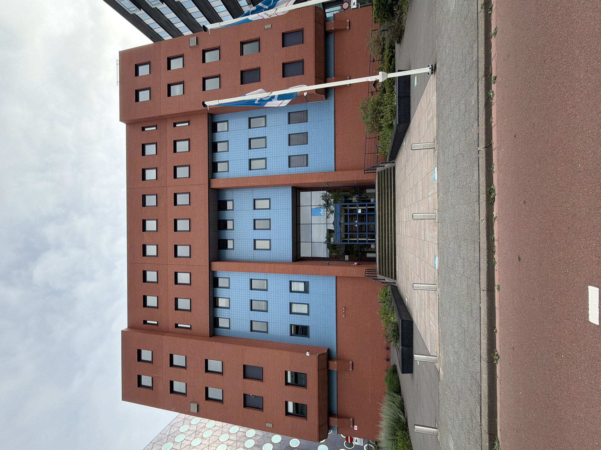 The image shows the CBR office building located in Rijswijk, Netherlands. The building is made of red brick and blue tiled sections and has many windows. In front of the building, there are stairs leading to the entrance, a paved area with bollards, and some landscaping. Two flags are visible on the right side of the building. The sky is overcast. There are no people visible in the image.