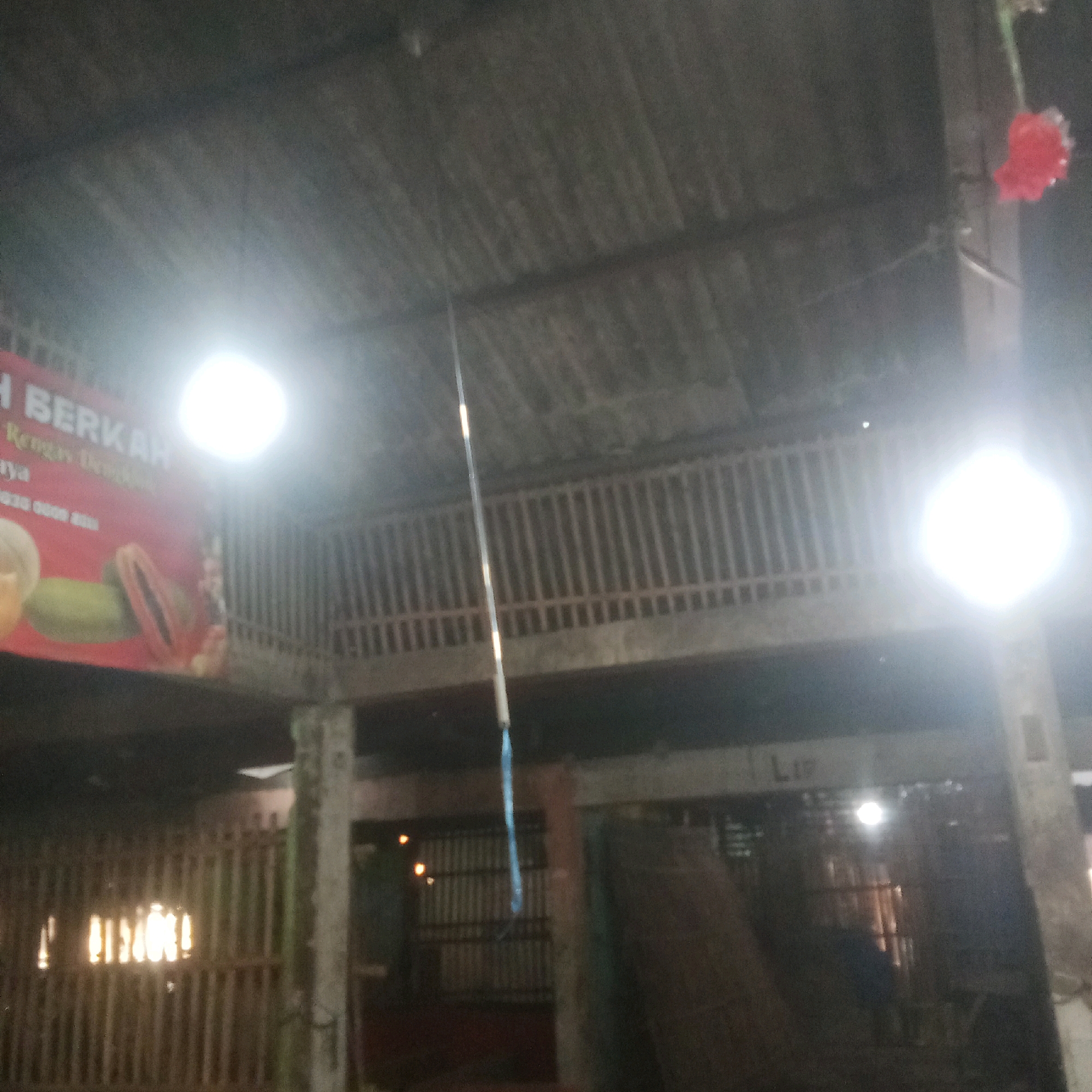 The image shows an indoor market or warehouse environment in Karawang, Indonesia. The scene is dimly lit with artificial lighting. In the background, there is a corrugated metal roof supported by wooden beams, and a wooden railing on an upper level. A red banner with white lettering is visible on the left, partially obscured by a bright light source. The banner appears to advertise fruits. Below, a wooden fence or partition runs horizontally, with more vertical wooden slats behind it. Some areas appear to be storage or market stalls. A long, thin object, possibly a pole with a blue string attached, hangs from the ceiling in the center of the image. There is a red decorative item hanging from a beam on the right.