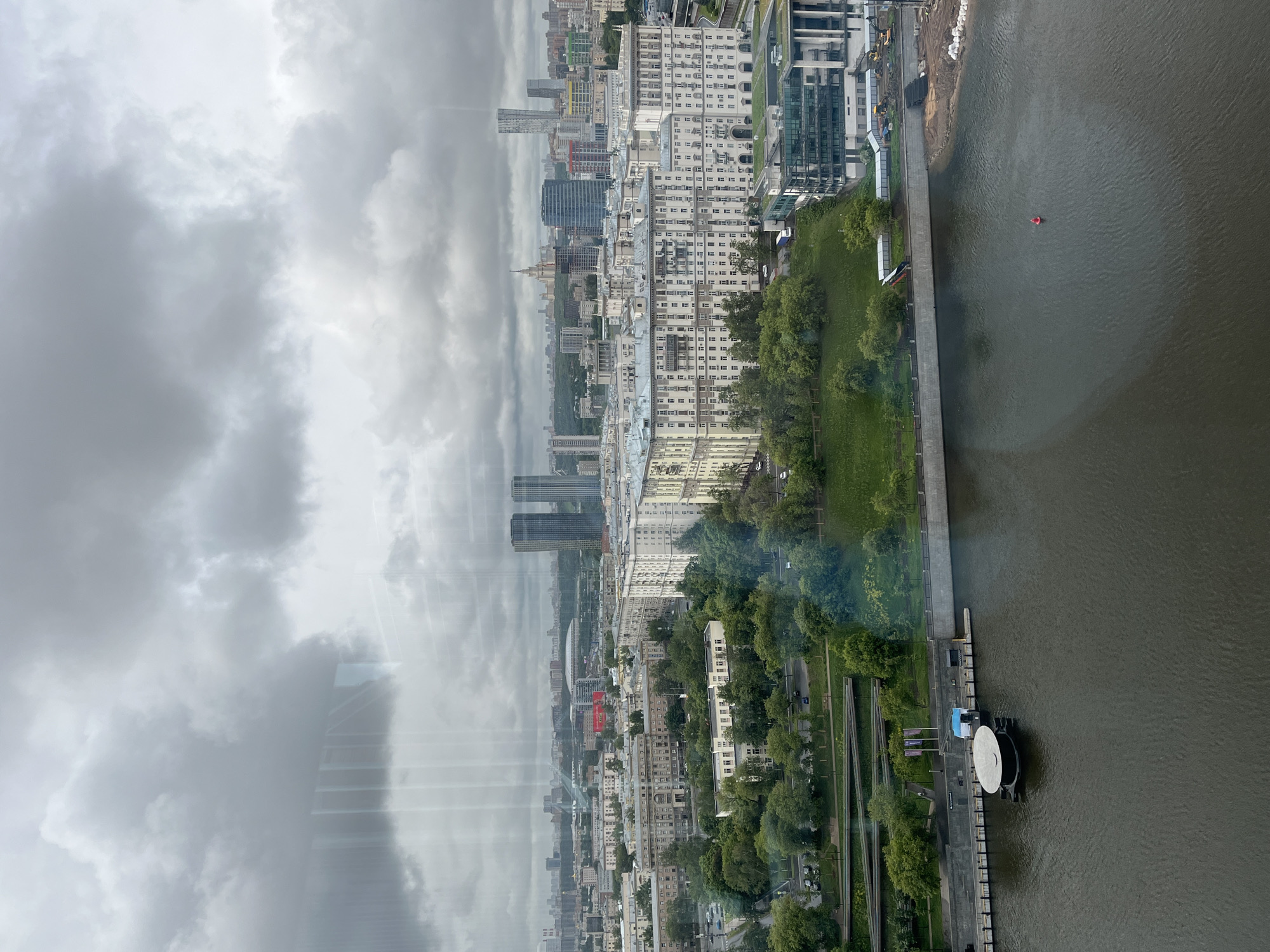 The image shows a cityscape taken from a high vantage point, potentially from inside a building looking out through a window. The city, likely Moscow based on the architecture, is overcast with a sky dominated by gray clouds. Below the clouds, the city unfolds, displaying a mix of older, light-colored apartment buildings and modern glass-clad skyscrapers. A river runs along the right side of the frame, with a long, straight embankment lined with green space and trees. Along the embankment, there's a distinctive circular structure, possibly a pier or a platform. In the river, there's a red buoy. The photograph provides an overview of the urban environment, emphasizing the juxtaposition of natural elements and urban development.