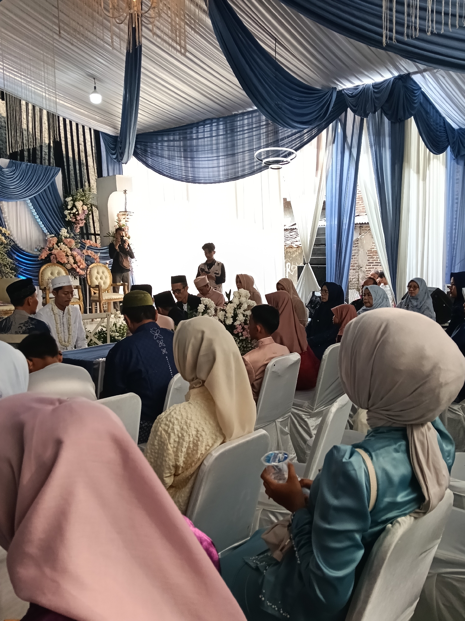 The image depicts a wedding ceremony taking place indoors, likely in Tegal Angus, Indonesia. The venue is decorated with draped white and blue fabric, creating a tent-like atmosphere. A chandelier hangs from the ceiling. In the foreground, several guests are seated, facing a central area where the ceremony is being held. Many of the women are wearing hijabs.
The main subjects of the ceremony are seated in the middle ground. A man in a white traditional outfit, likely the groom, is seated on the left, and another man in a similar outfit is to his right. There are other men and women gathered around them, some appearing to be officiants or family members. One person is holding a camera and taking pictures. White chairs are arranged for the guests, and a table with what appears to be wedding decorations and flowers is visible. The overall scene suggests a formal, traditional wedding event.