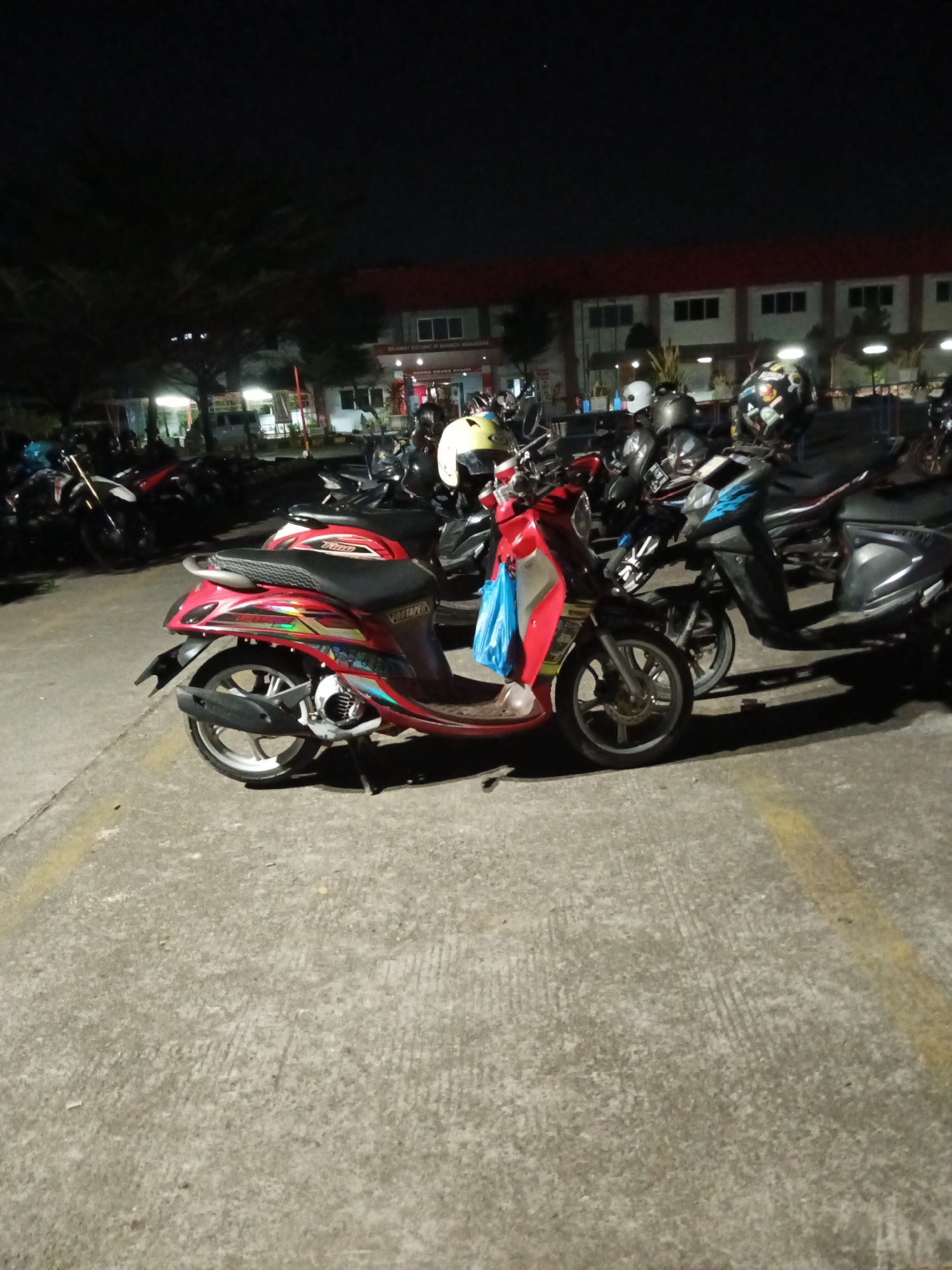A low-angle, medium shot depicts a dark, outdoor parking lot at night, likely in Makassar, Indonesia, with numerous parked motor scooters. The primary focus is a red scooter in the foreground, positioned on a concrete surface marked with yellow lines. This scooter features a black seat, a silver rim on the rear wheel, and intricate colorful decals along its side. A bright blue plastic bag hangs from its handlebars. Behind the red scooter, other scooters are visible, some with helmets on their seats, including a white helmet and a black helmet with a sticker design. In the background, a building with red trim and multiple windows is illuminated, suggesting it is a commercial or institutional establishment. The scene is dimly lit by artificial lights, casting shadows and creating a somber atmosphere. The overall environment appears to be a secured parking area, possibly belonging to a business or public facility.