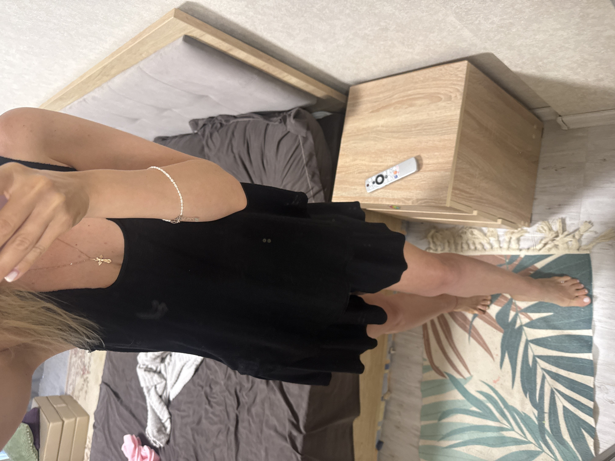 A woman stands in a bedroom. She is wearing a black tank top and black shorts. She has a gold necklace with a cross pendant and a pearl bracelet on her left wrist. Her toenails are painted pink. The bed has a gray headboard and is covered with a dark gray comforter. A nightstand to the right of the bed holds a white remote control. A rug with a palm leaf pattern is on the floor in front of the bed. There are stacks of boxes and some clothing on the floor to the left of the bed. The walls are light gray and textured.