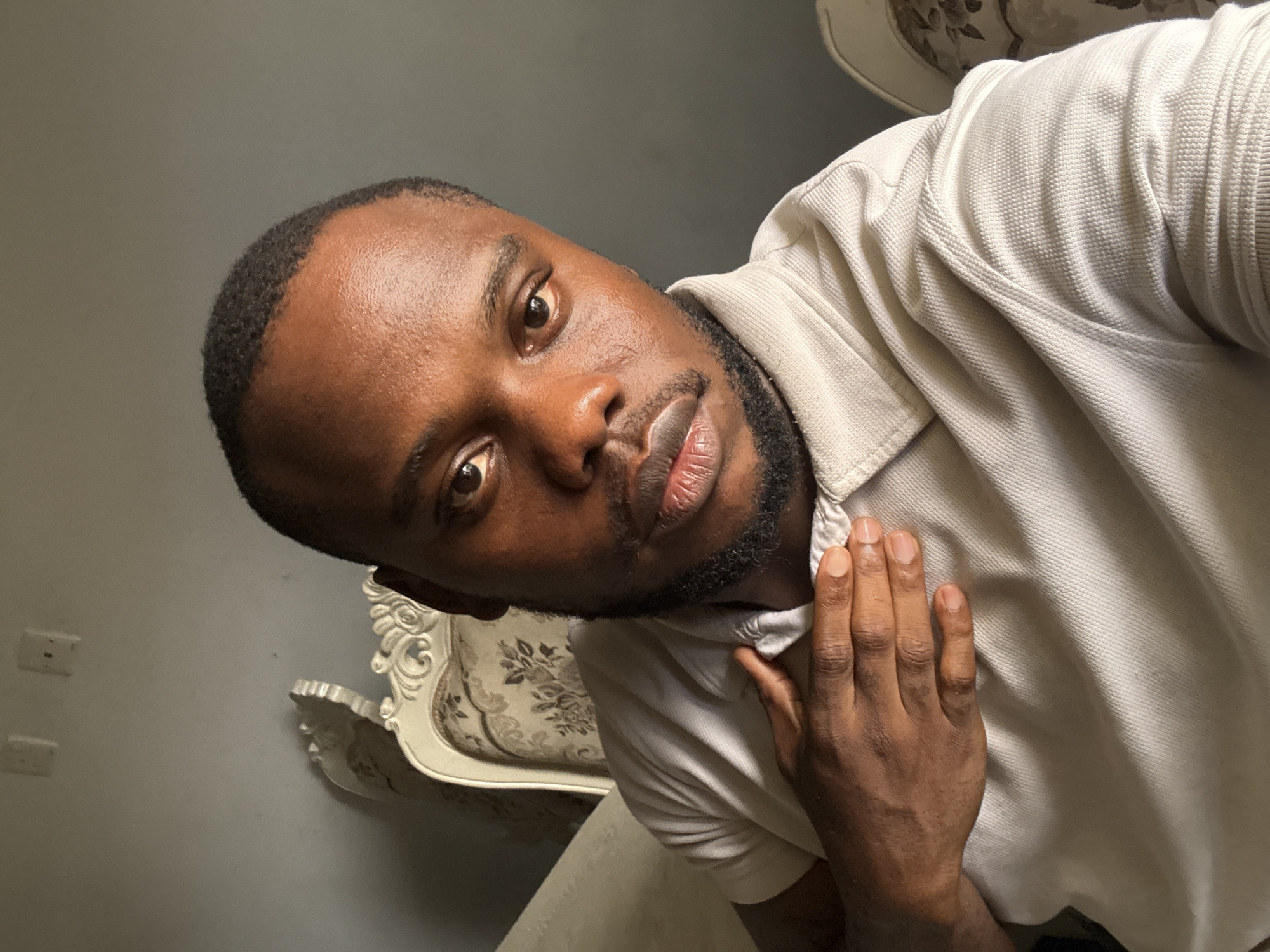 A close-up, low-angle shot depicts a dark-skinned man with short, black hair and a beard. He is wearing a light-colored polo shirt. His right hand is raised near his chest. In the background, a white, ornate chair is partially visible. The setting appears to be indoors, with a plain wall and electrical outlets also in the background. There is no specific information available to place the location in Iddo Tudunwada, Nigeria.